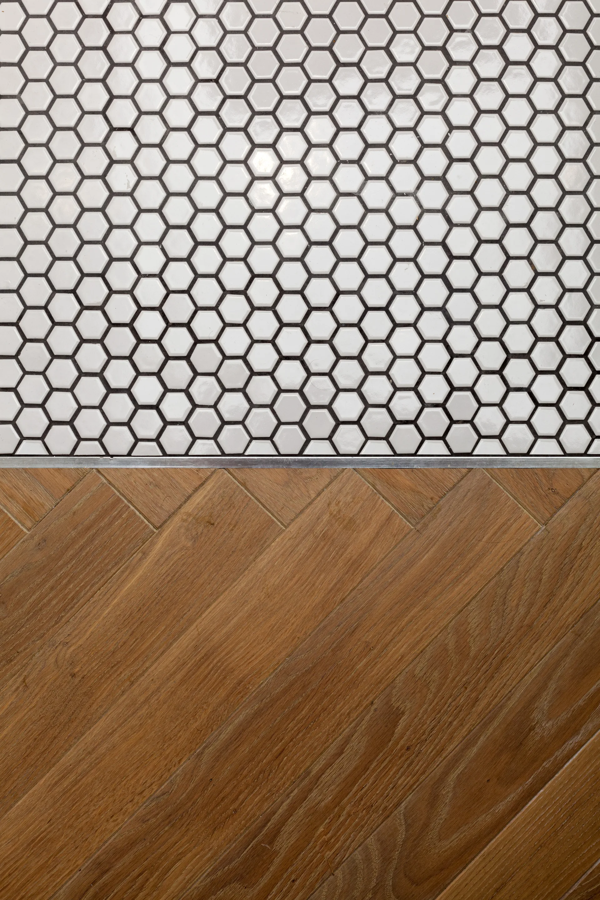 A top-down view shows white hexagonal tile with dark grout meeting diagonal wood planks at a slim metal transition strip.