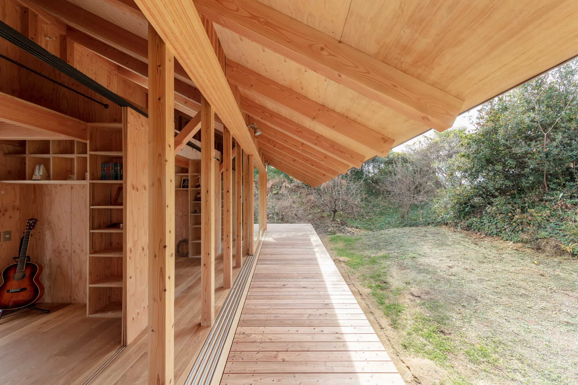 A covered wooden deck with a deeply sloped roof extends from a light plywood interior featuring built-in shelving and an acoustic guitar.