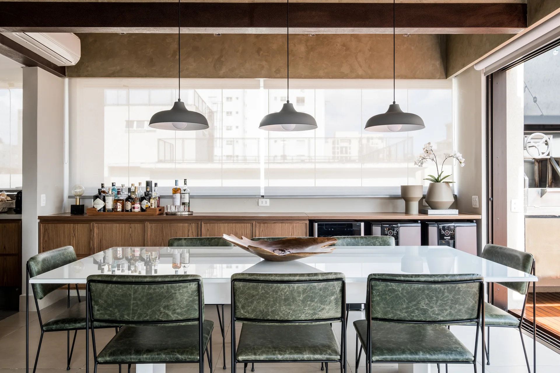 A glossy white dining table surrounded by green leather-style chairs under three dark dome pendant lights.