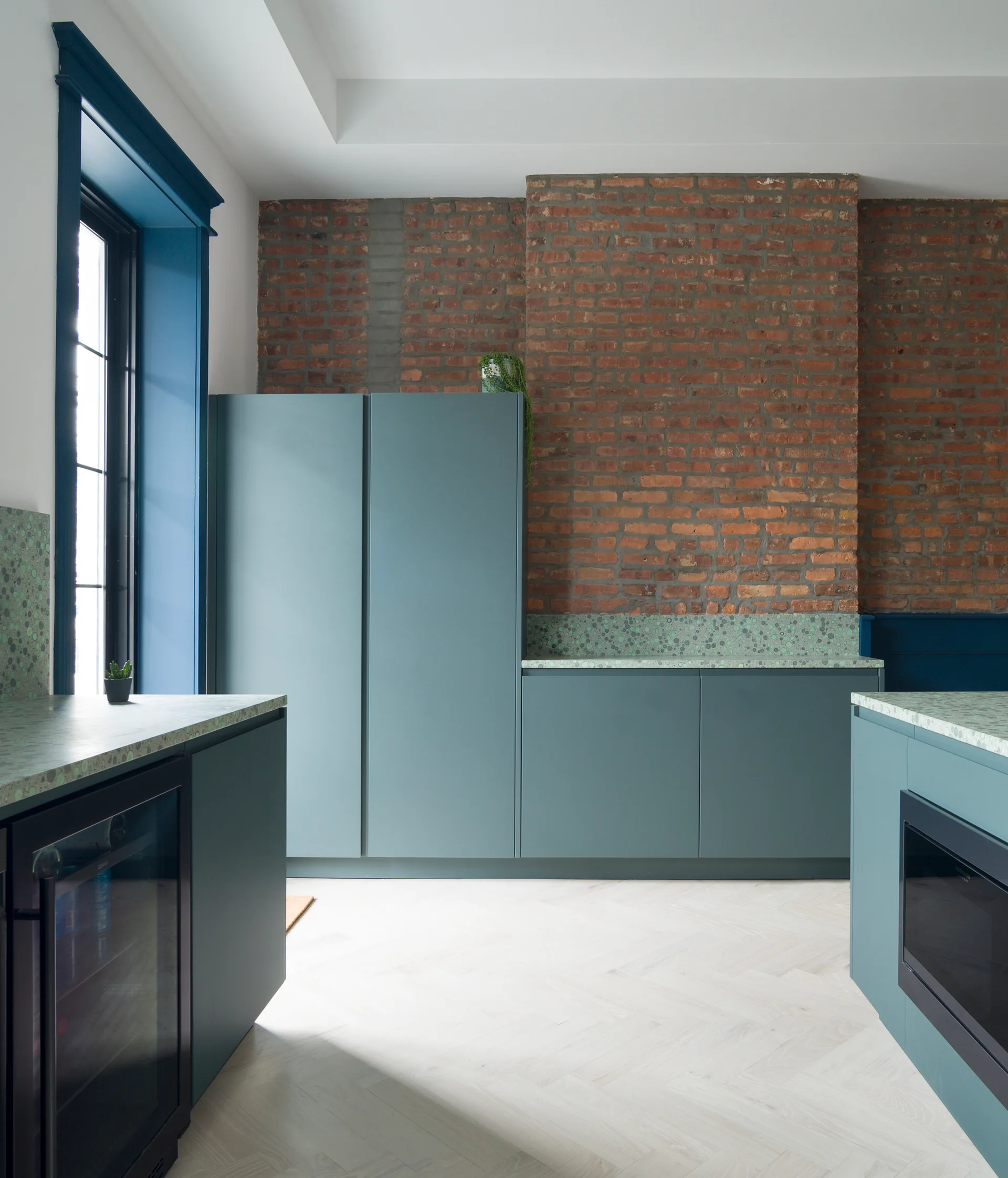A modern kitchen with matte blue-green cabinets, green terrazzo countertops, an exposed brick wall, and light herringbone floors.