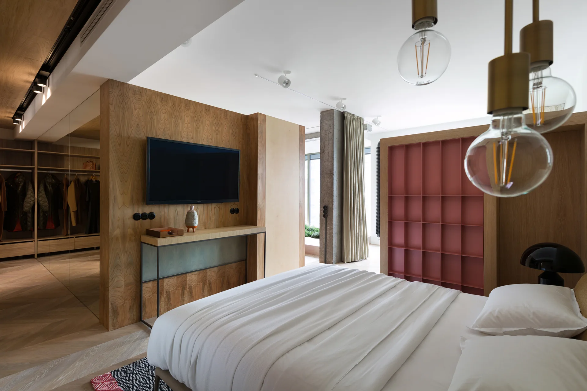 A modern bedroom features a bed with white linens, a wood-paneled partition wall with a mounted TV, and an open walk-in closet.