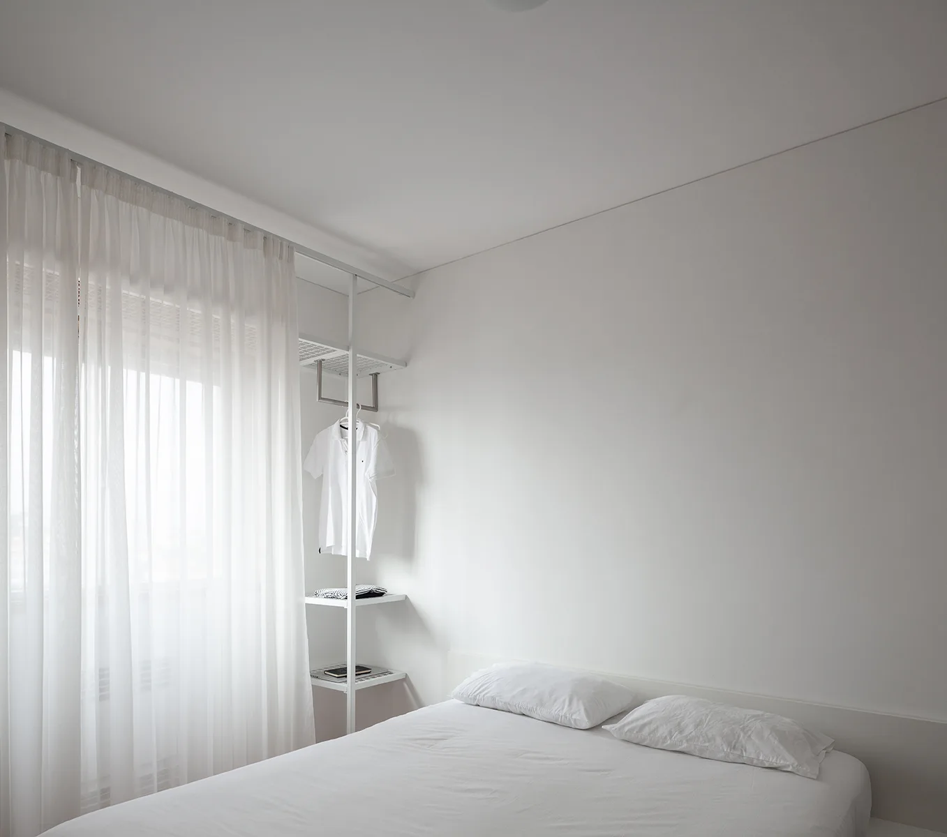 A minimalist white bedroom featuring a bed with white linens next to a white metal open shelving unit and sheer window curtains.
