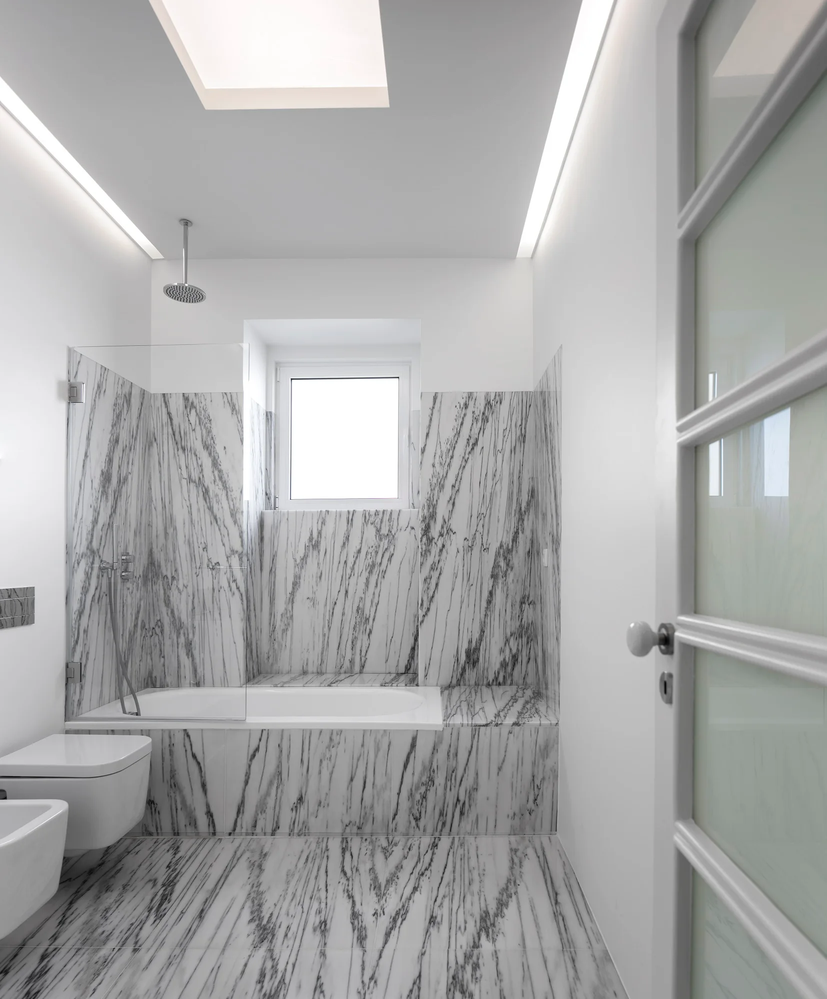 A bright minimalist bathroom featuring heavily veined white marble flooring, a marble-clad bathtub surround, a skylight, and wall-mounted toilet.