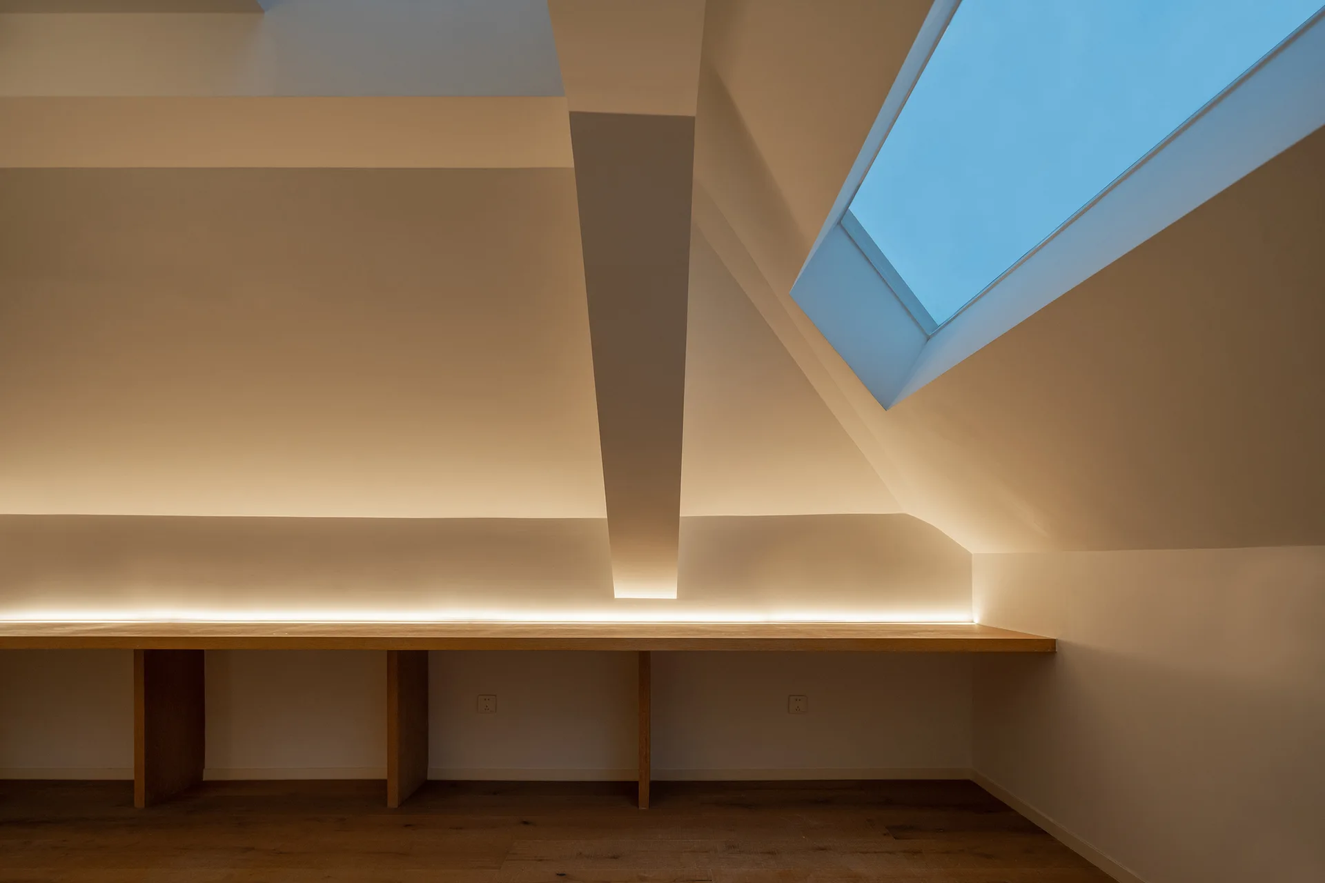 A minimalist attic room featuring a long built-in wooden desk, a slanted ceiling with a skylight, and integrated LED strip lighting.