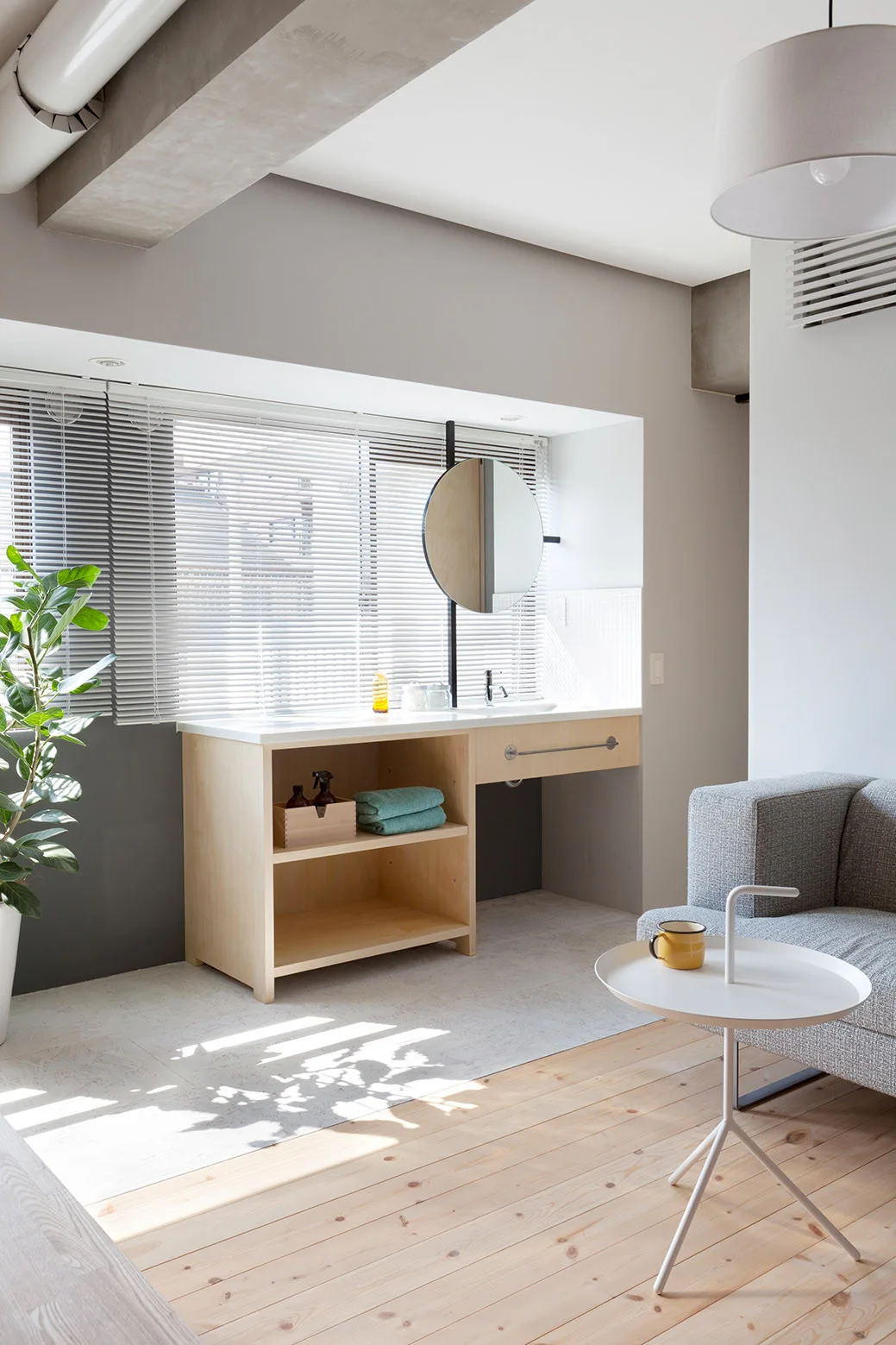 A light wood vanity with a round mirror sits under a window with blinds, next to a grey sofa on mixed wood and tile floors.