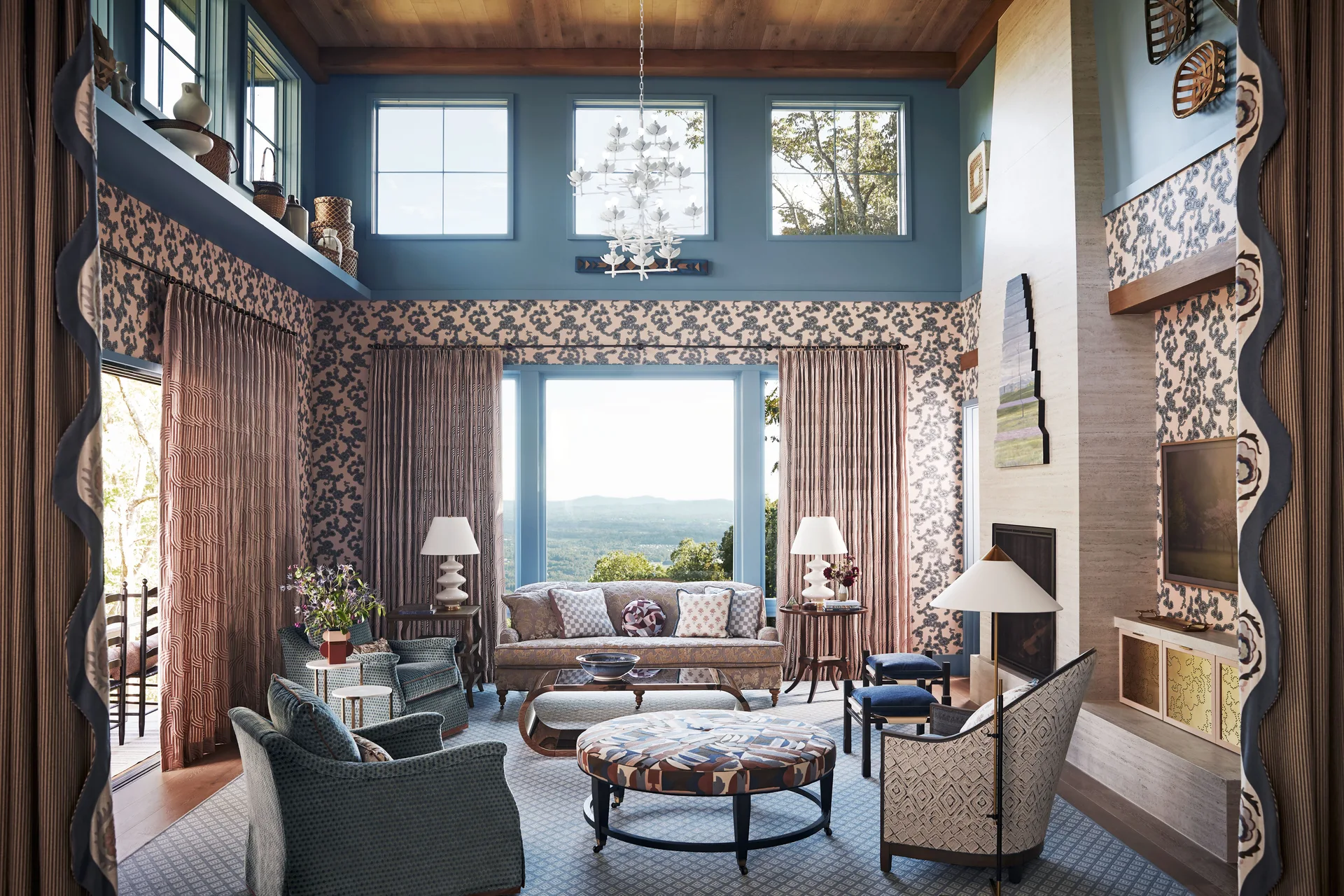 A high-ceilinged living room features blue patterned wallpaper, a stone fireplace, patterned seating, and a large window with mountain views.