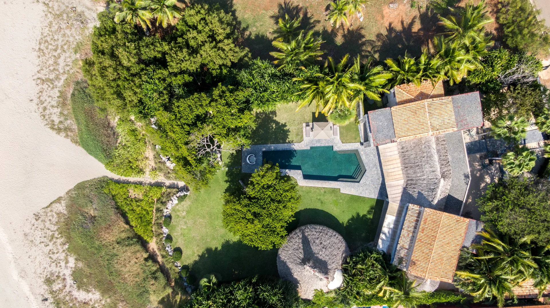 An aerial view of a beachfront property featuring a central swimming pool, stone patio, green lawns, and structures with terracotta and thatched roofs.