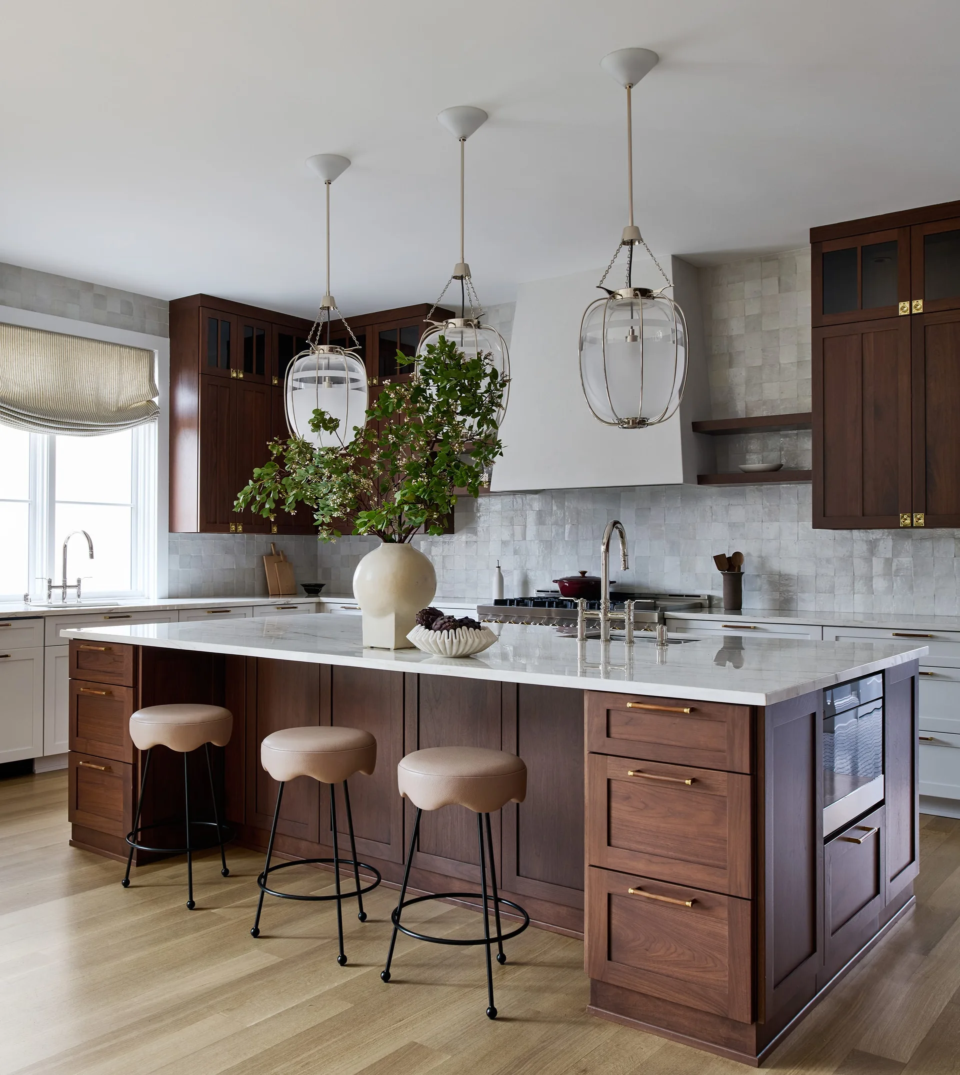 A transitional kitchen featuring dark wood cabinetry, a large marble-topped island with three wavy-seat bar stools, and glass lantern pendant lights.