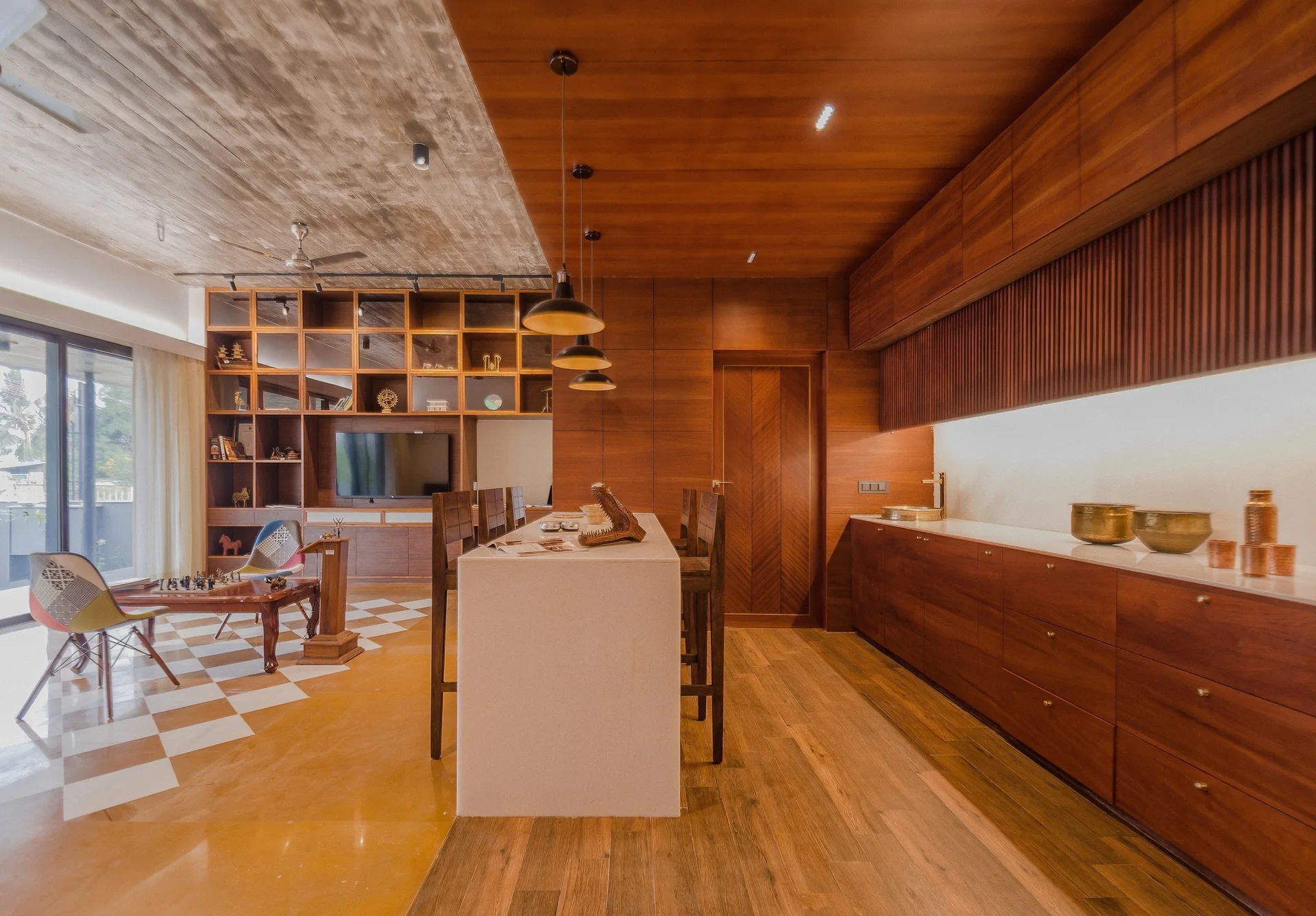 A warm wood kitchen featuring a stone island and cabinetry, situated next to a living area with a checkered floor and exposed concrete ceiling.