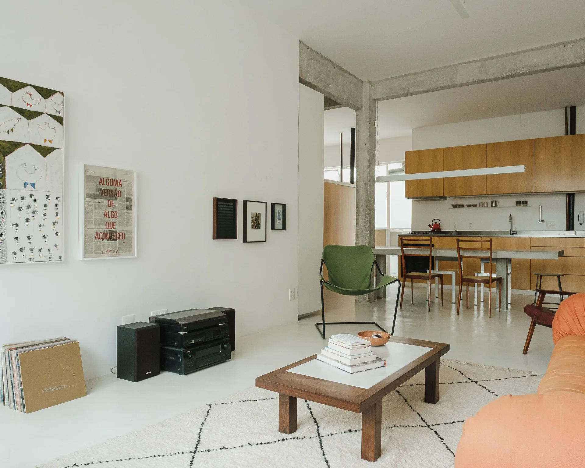 An open concept living room featuring an orange sofa, a low wooden coffee table, a green accent chair, and an exposed concrete ceiling beam.