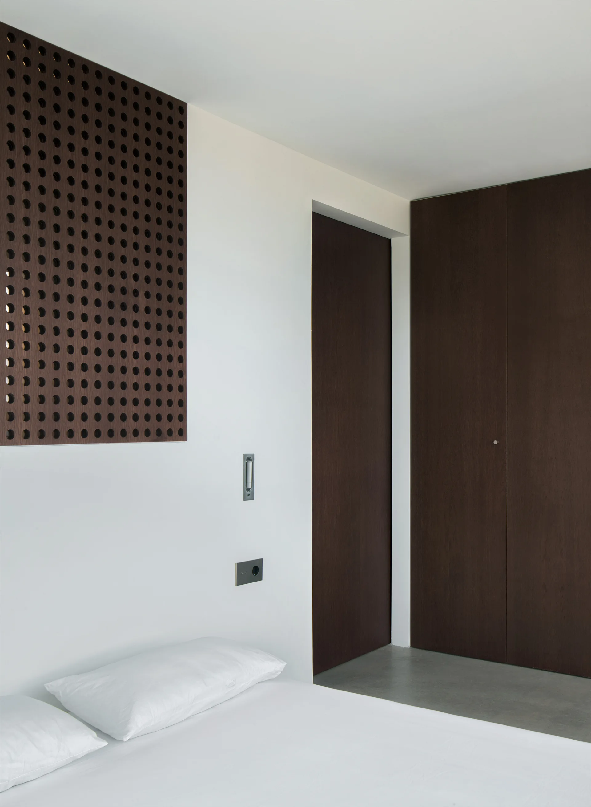 A minimalist bedroom featuring white bed pillows below a dark brown perforated wood panel next to tall flush closet doors.