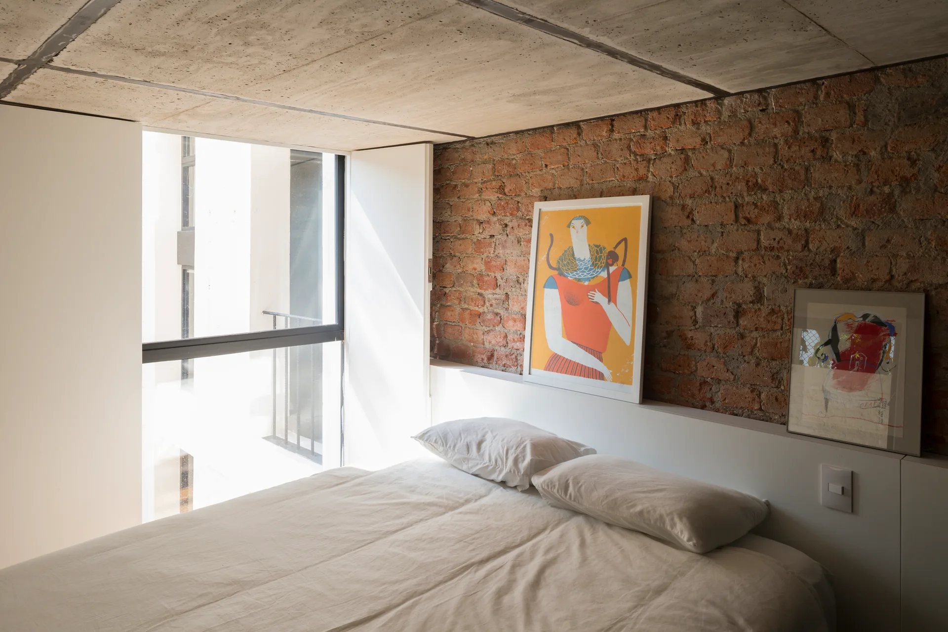 A bed with white pillows is positioned against a built-in headboard ledge in a bedroom with an exposed brick wall and raw concrete ceiling.