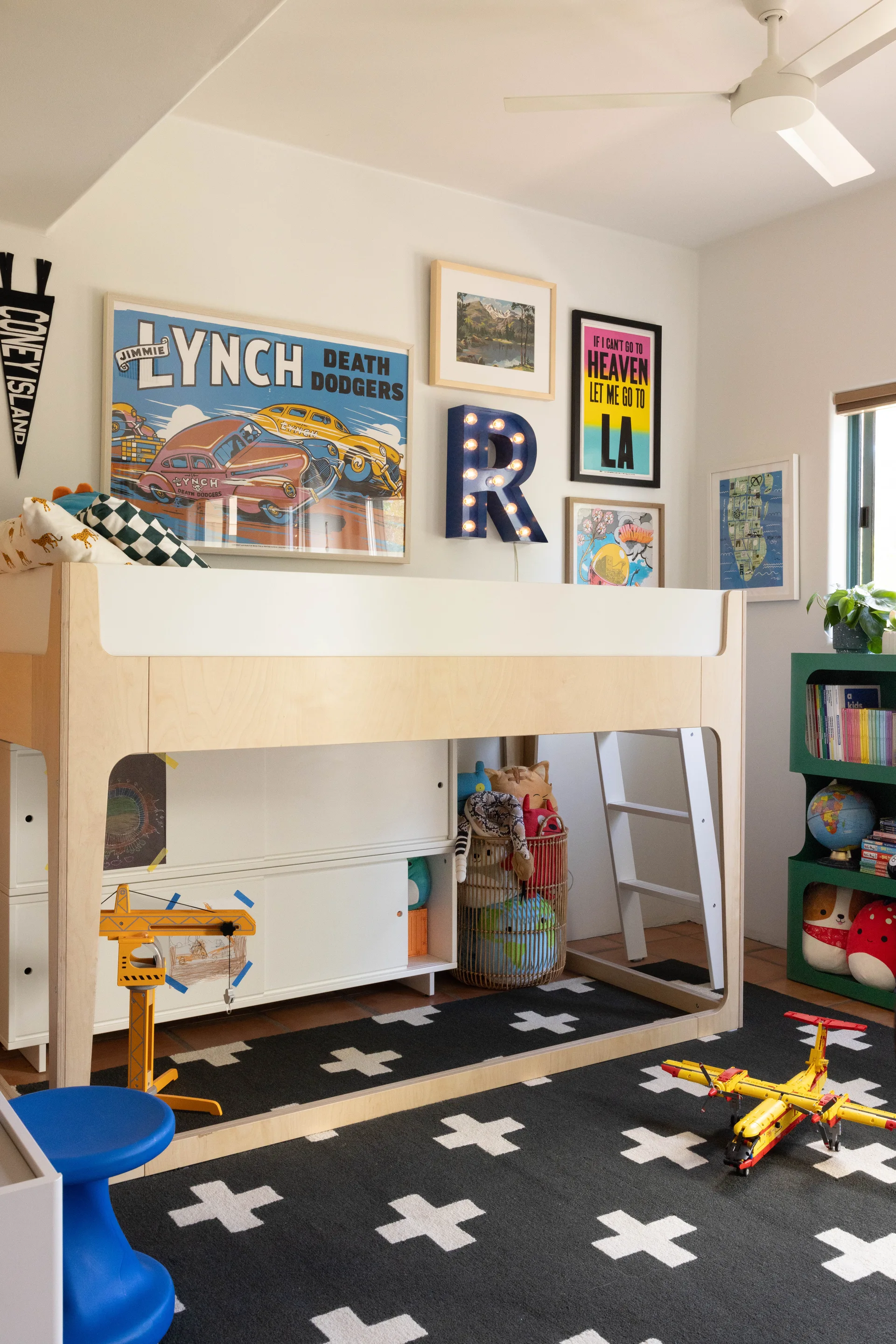 A light wood loft bed sits on a black and white cross-patterned rug in a kids' bedroom decorated with colorful wall art and a green bookshelf.