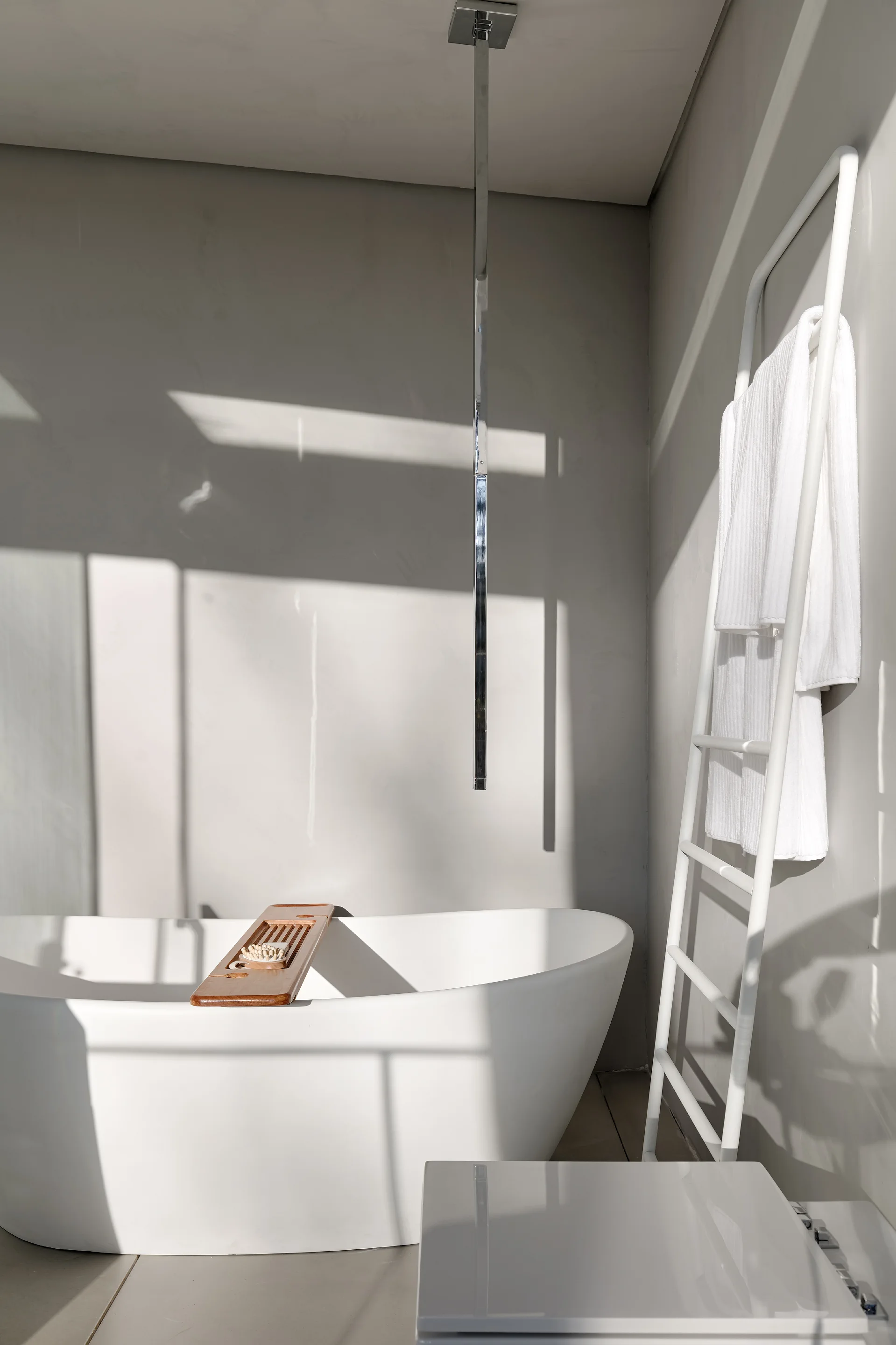 A white freestanding bathtub with a wooden tray sits in a minimalist grey bathroom next to a white leaning towel ladder.