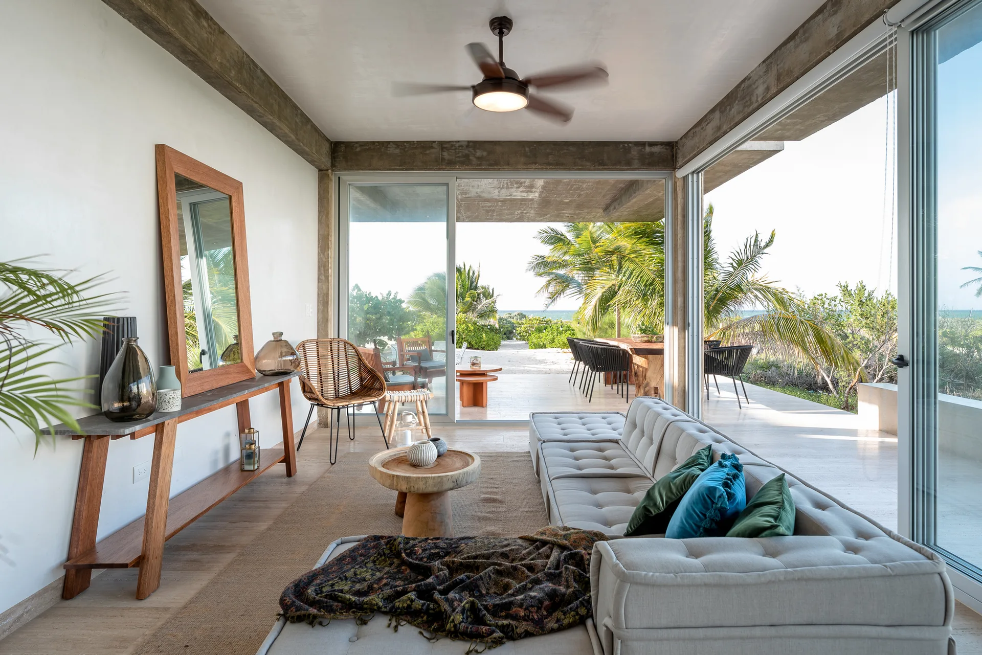 A bright living room with a tufted sectional sofa, a wooden console table with a large mirror, and open sliding doors leading to an outdoor terrace.