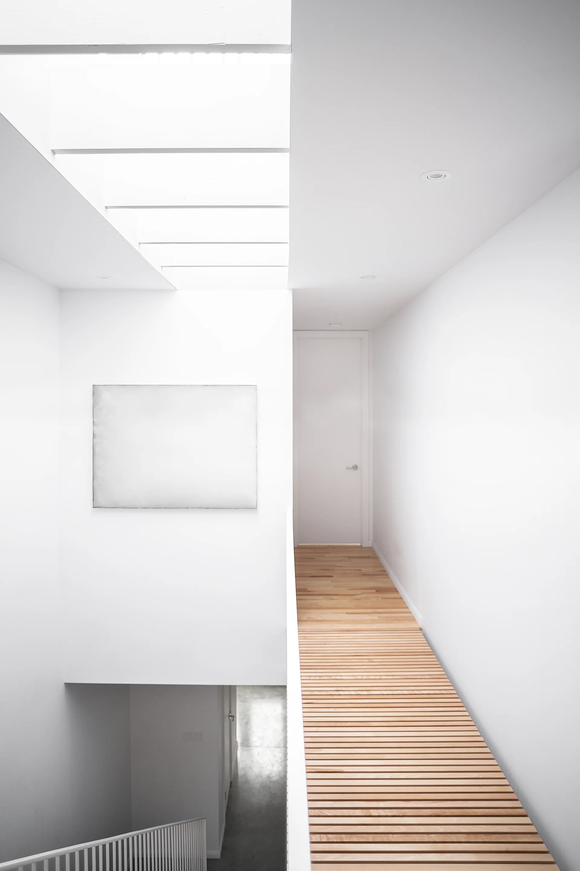 A minimalist hallway features a slatted wood walkway, a large overhead skylight, white walls, and a staircase railing below.