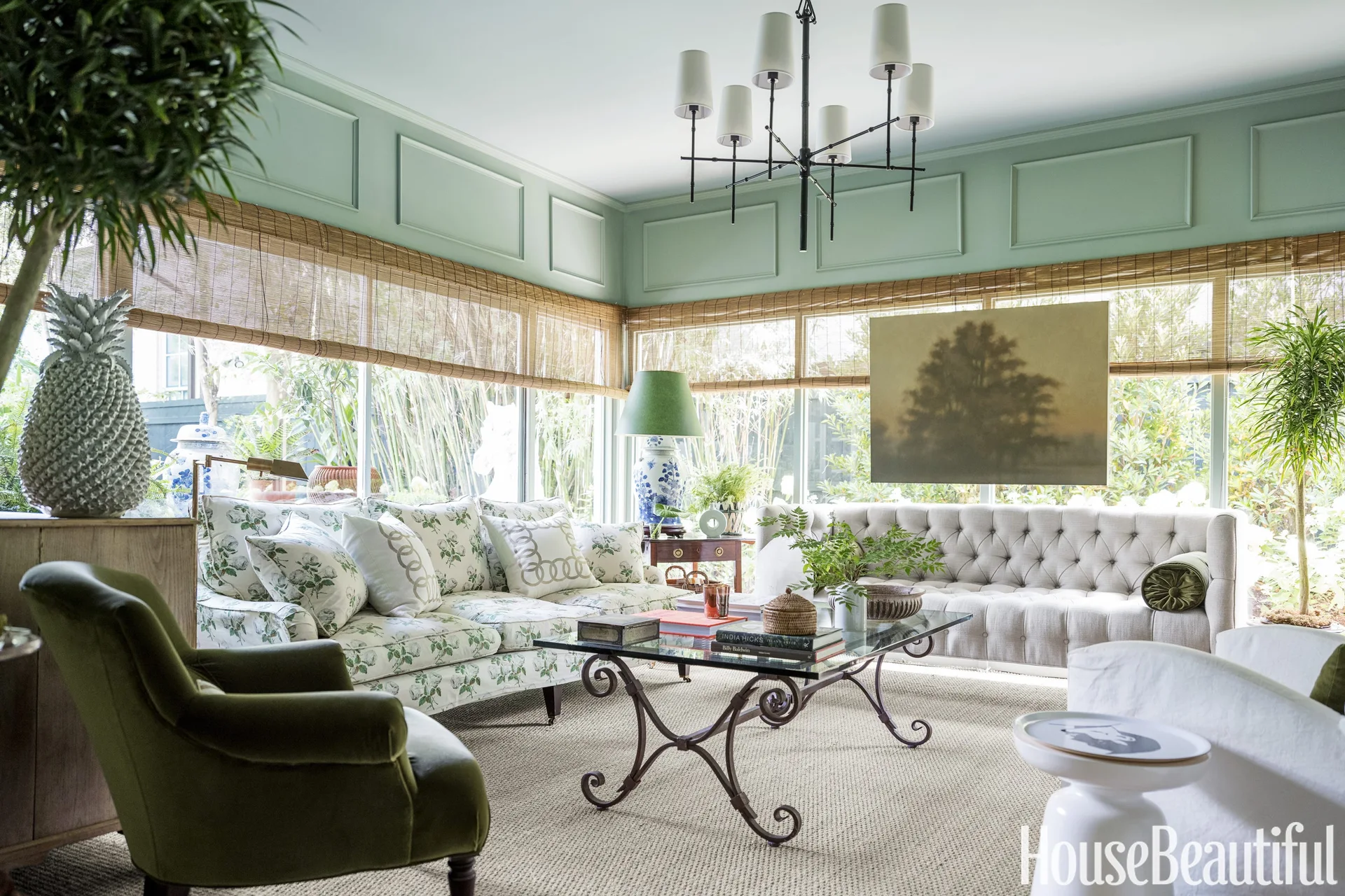 A sunlit living room featuring sage green paneled walls, a floral print sofa, a tufted cream sofa, and woven bamboo shades over large windows.