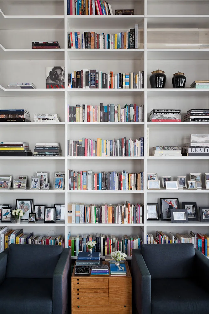 Two dark blue armchairs flank a small wood chest of drawers in front of a massive white built-in bookcase filled with books and pictures.