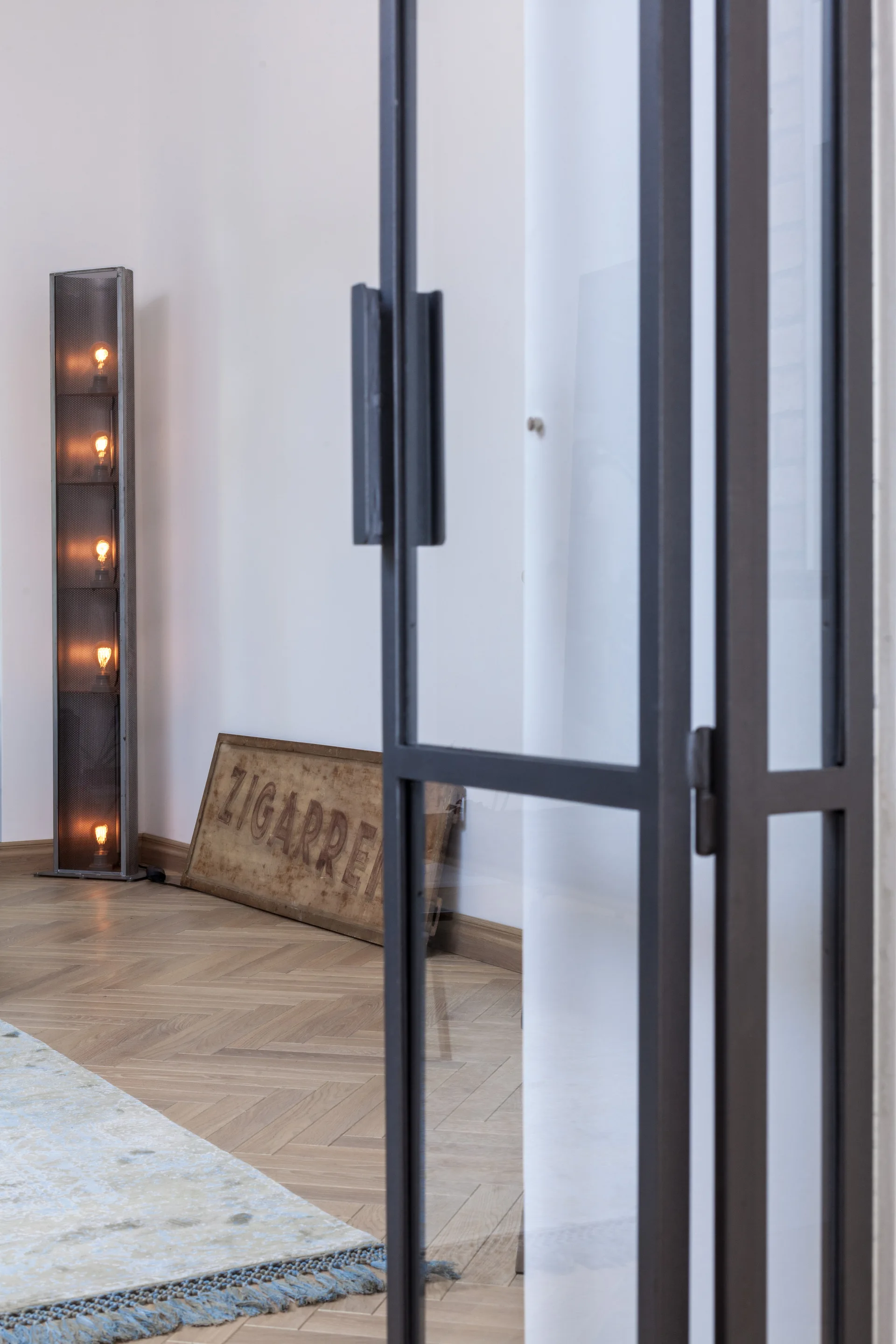 A black metal-framed glass door opens to a room with light wood herringbone flooring, a tall mesh floor lamp, and a leaning vintage wooden sign.