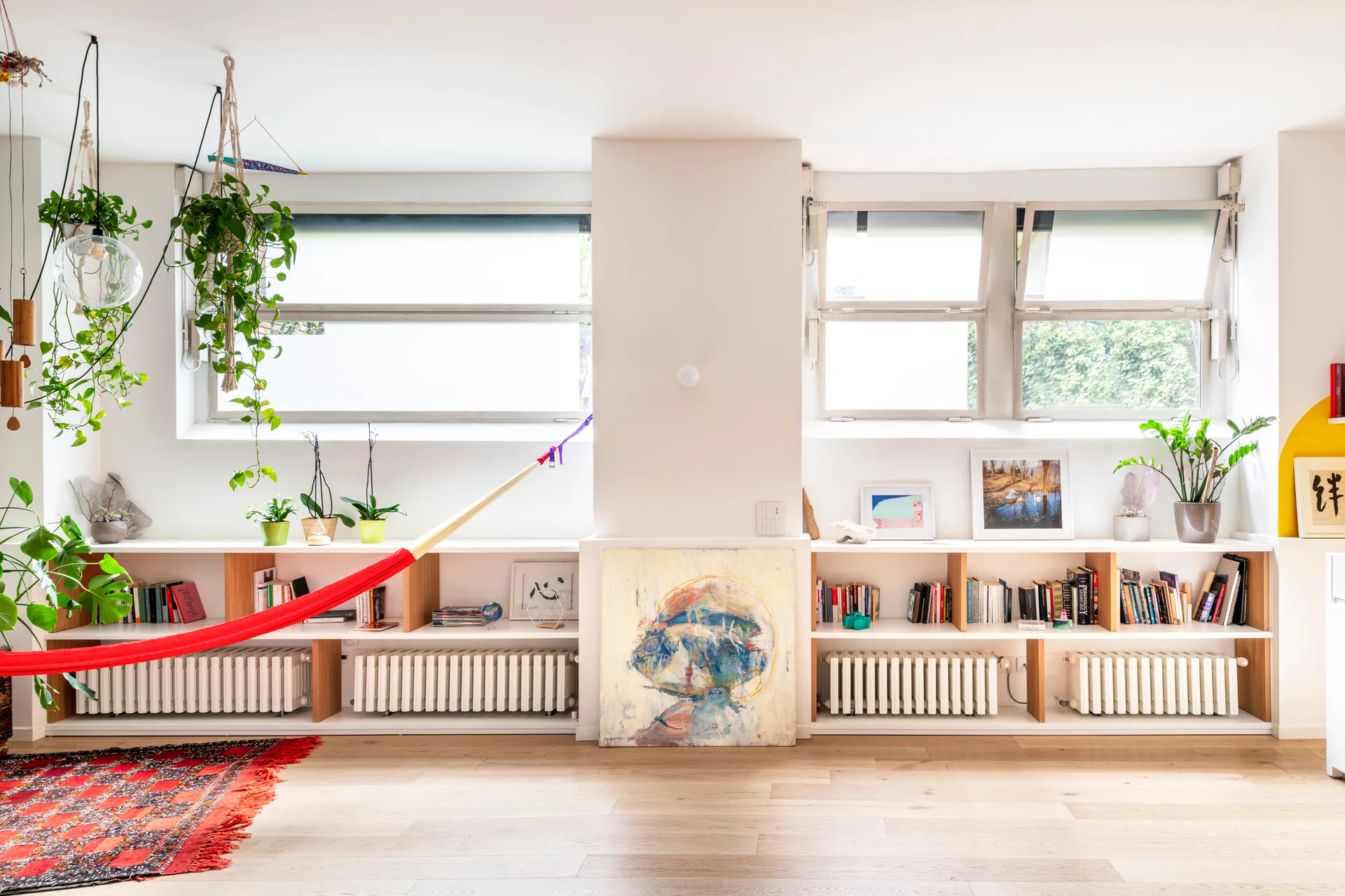 A bright room with a long built-in white bookshelf featuring integrated radiators, potted plants, books, and a vibrant red hammock suspended in front.