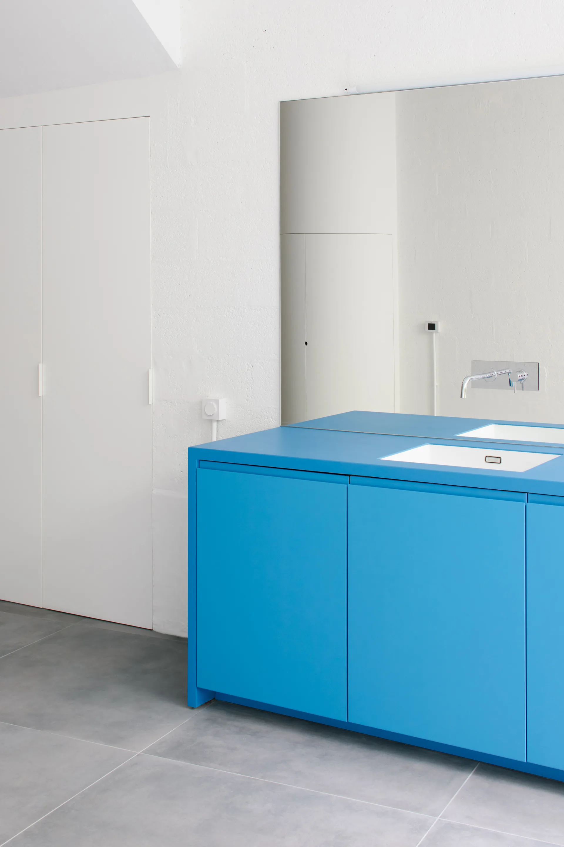 A minimalist bathroom featuring a bright blue cabinet vanity with an integrated white sink, a large frameless mirror, and grey tile floors.