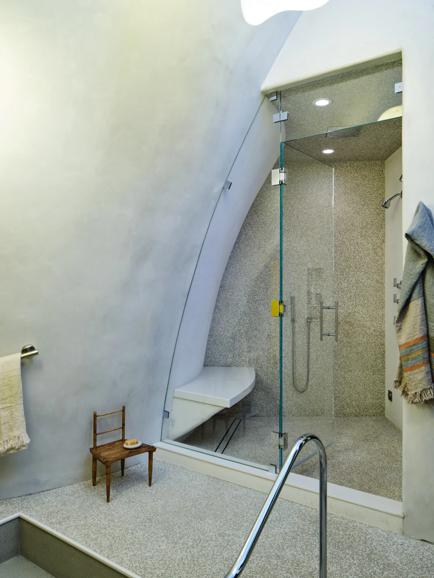 A contemporary bathroom features a glass shower enclosure with gray mosaic tile, a white built-in bench, and a smooth curved plaster wall.