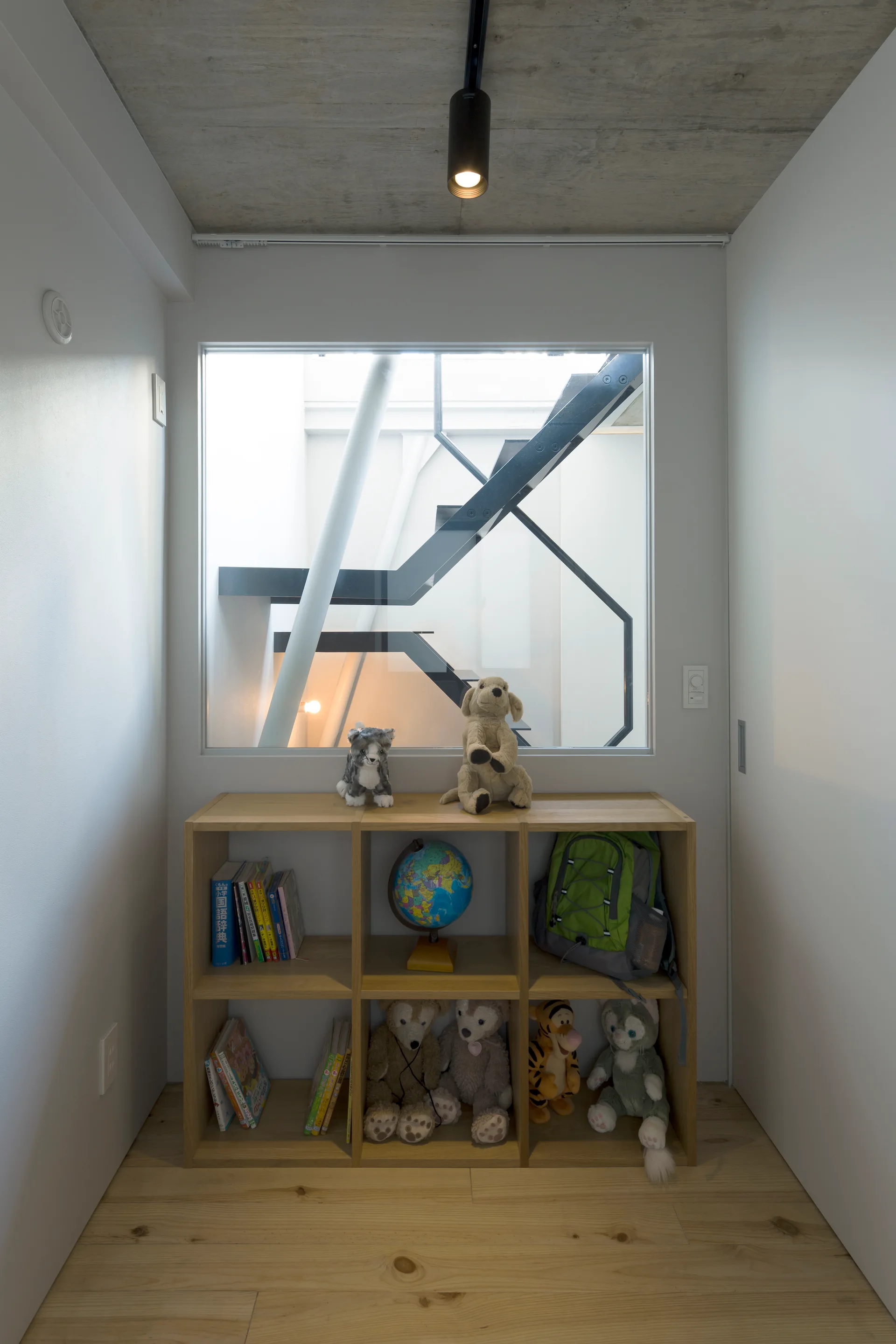 A wooden cubby bookshelf filled with books and stuffed toys sits below an interior window looking out onto a black steel staircase.