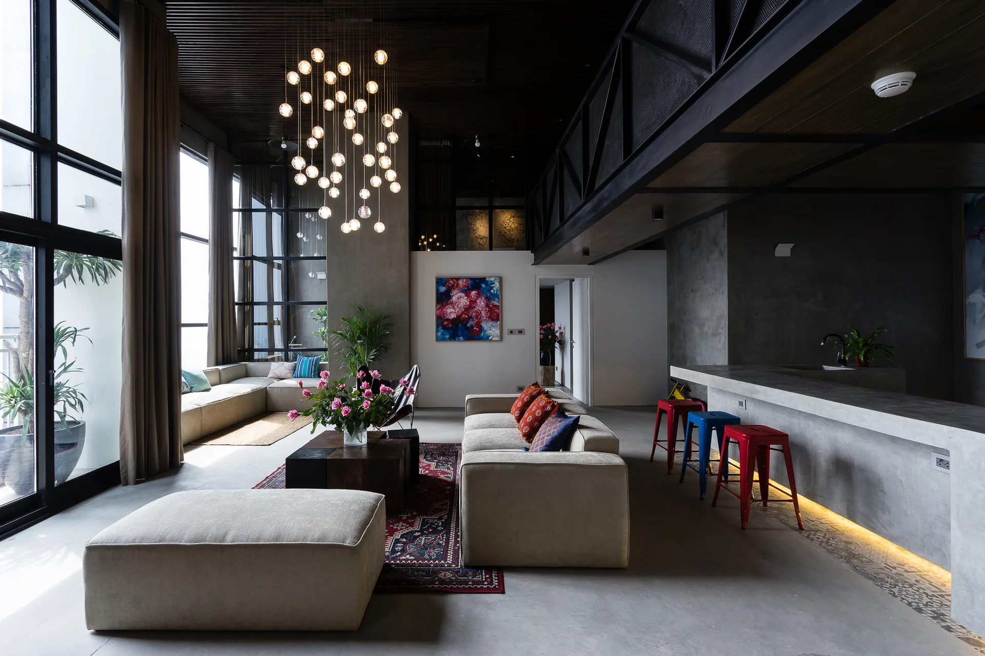 An industrial living room with concrete floors, beige sofas, a bubble chandelier, and a concrete kitchen counter with colorful metal stools.