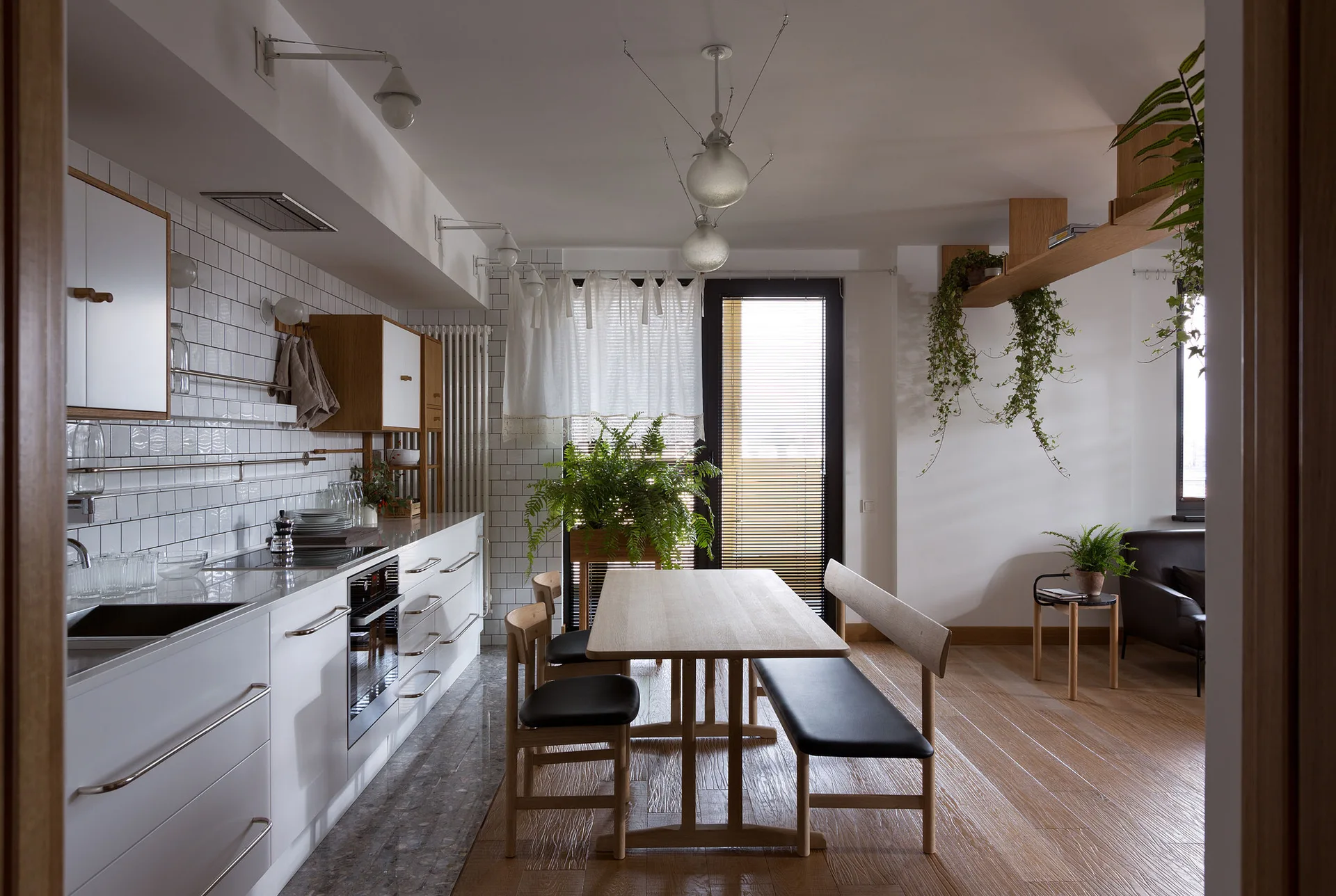 A Scandinavian kitchen and dining area features white cabinetry, a wood dining table with benches, and a white subway tile backsplash wall.