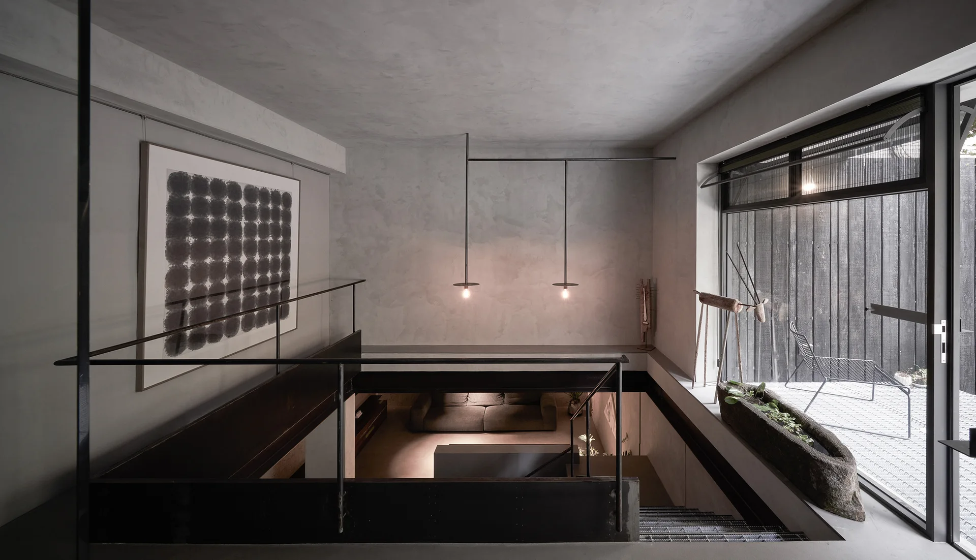 A concrete stairwell with black metal railings leads to a lower-level living area, featuring minimalist pendant lights and textured grey walls.