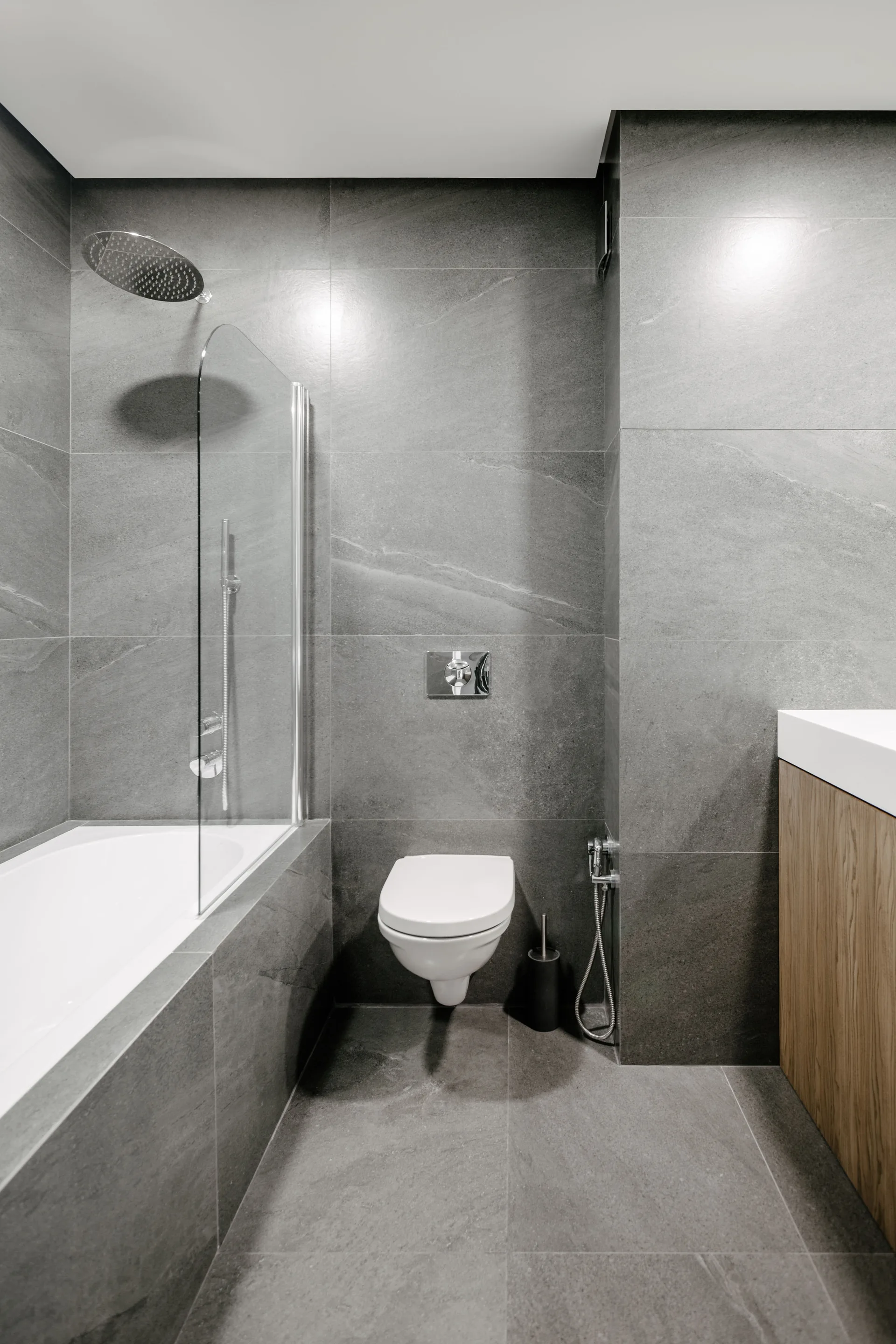 A modern bathroom clad in grey stone tiles features a wall-mounted toilet, a bathtub with a glass screen, and a wood vanity cabinet.