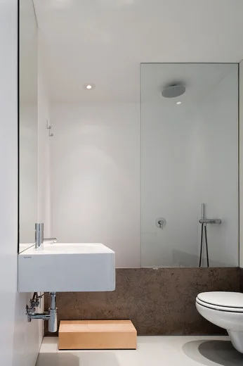A minimalist white bathroom features a wall-mounted rectangular sink, a wall-hung toilet, and a glass shower partition beside a dark brown stone band.