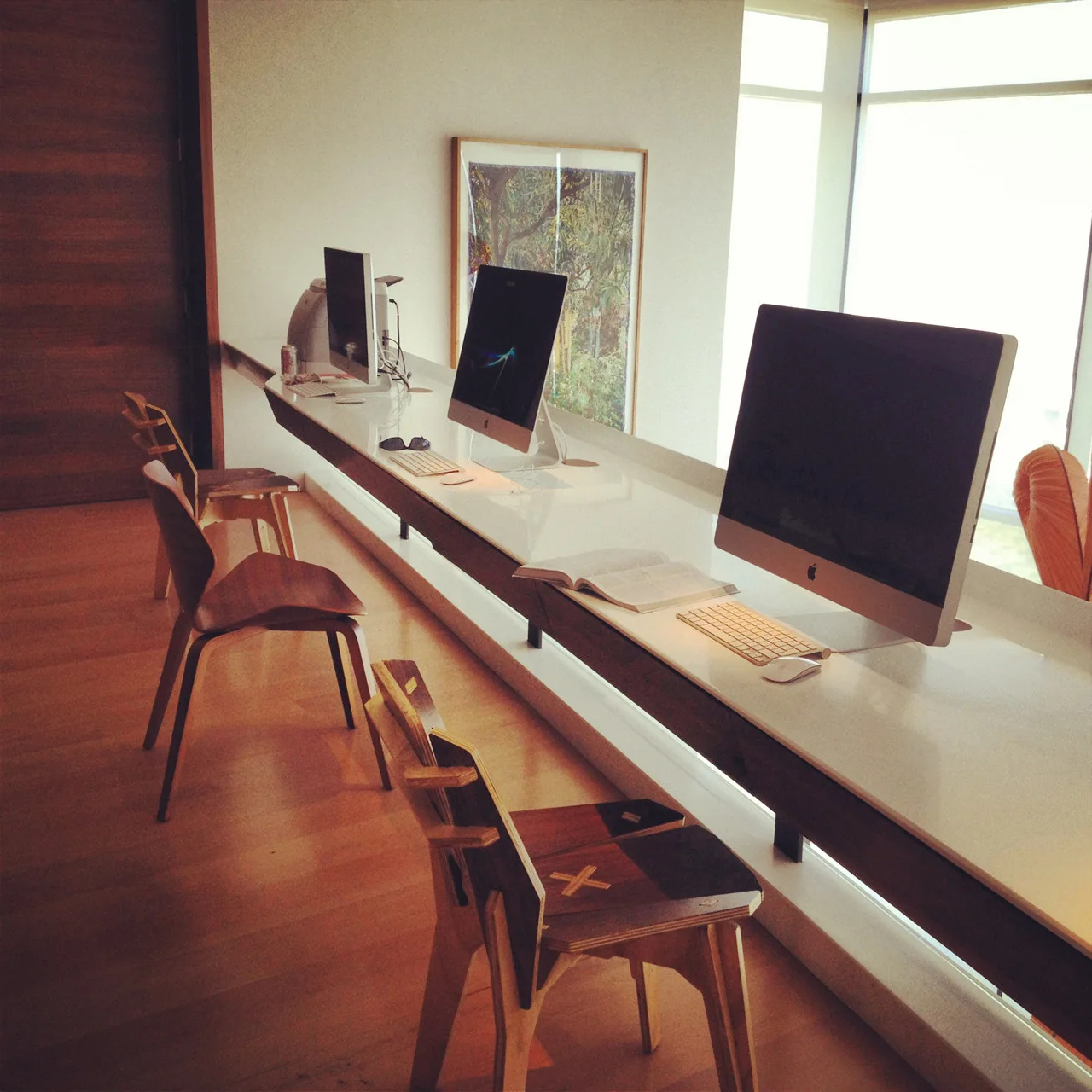 A long, built-in white floating desk in a home office is lined with three computer setups and several wooden chairs on wood flooring.