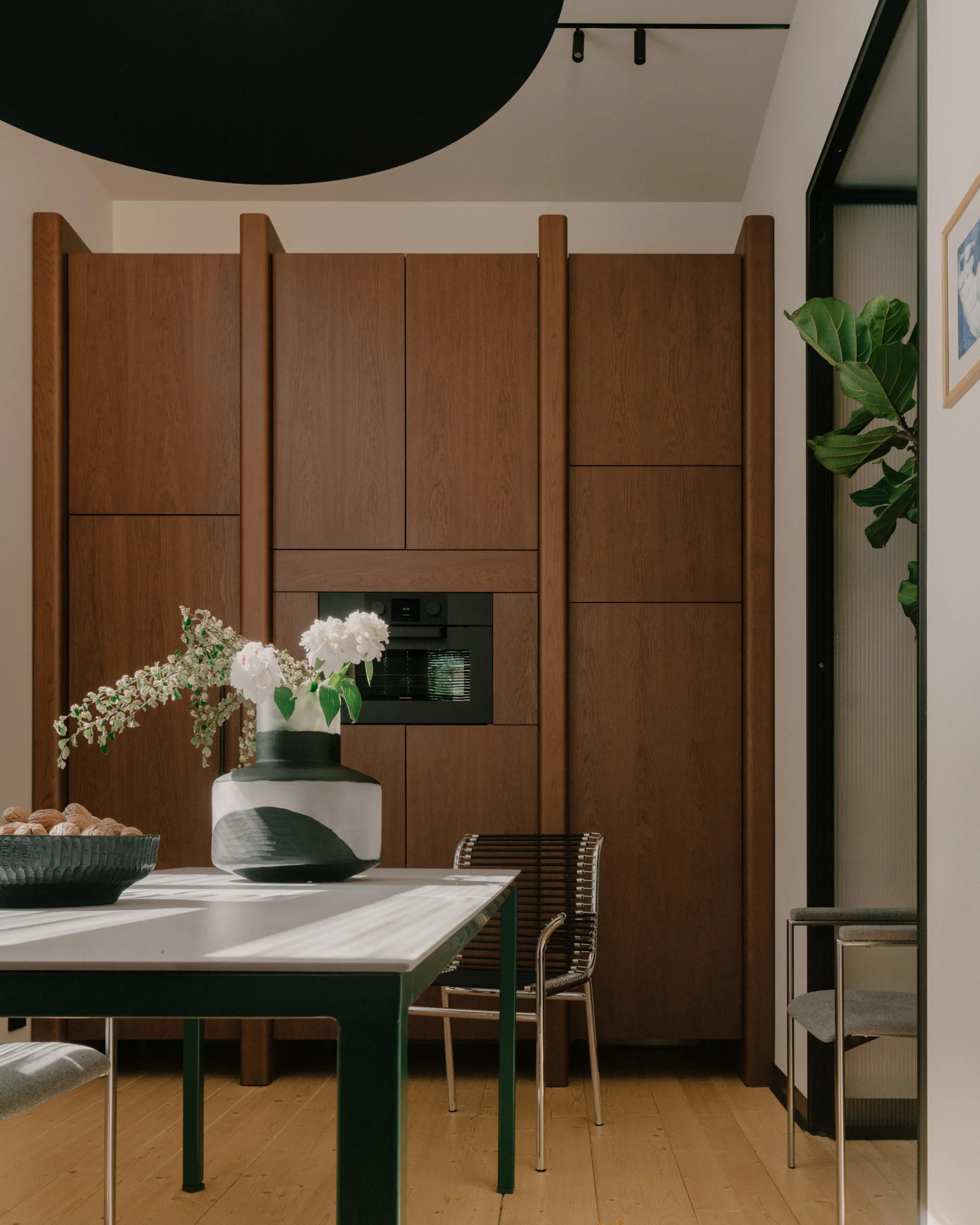 A dining room features a green-framed table with a graphic ceramic vase, a cord-seat chair, and a wall of built-in wooden cabinets.