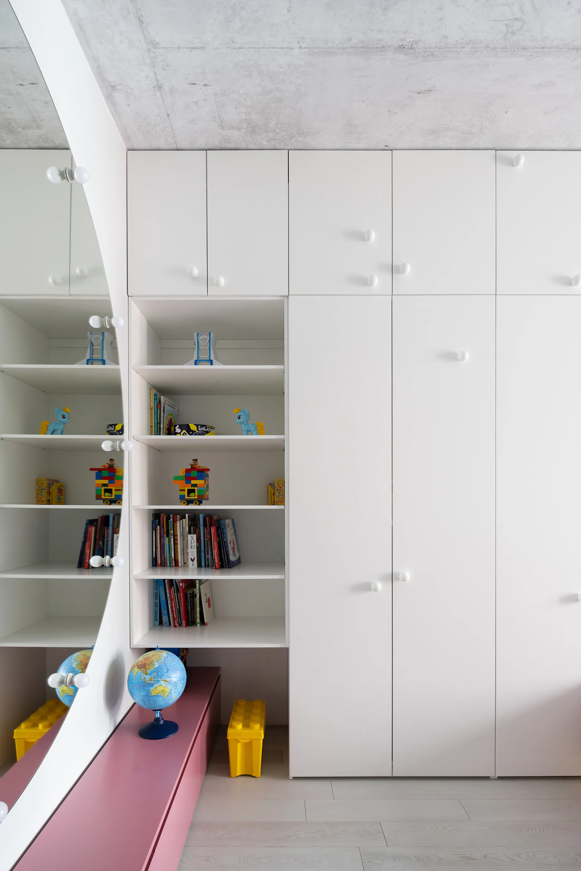 A white built-in wardrobe in a child's bedroom features open shelves with toys, a vertical mirror with lights, and a low pink bench.