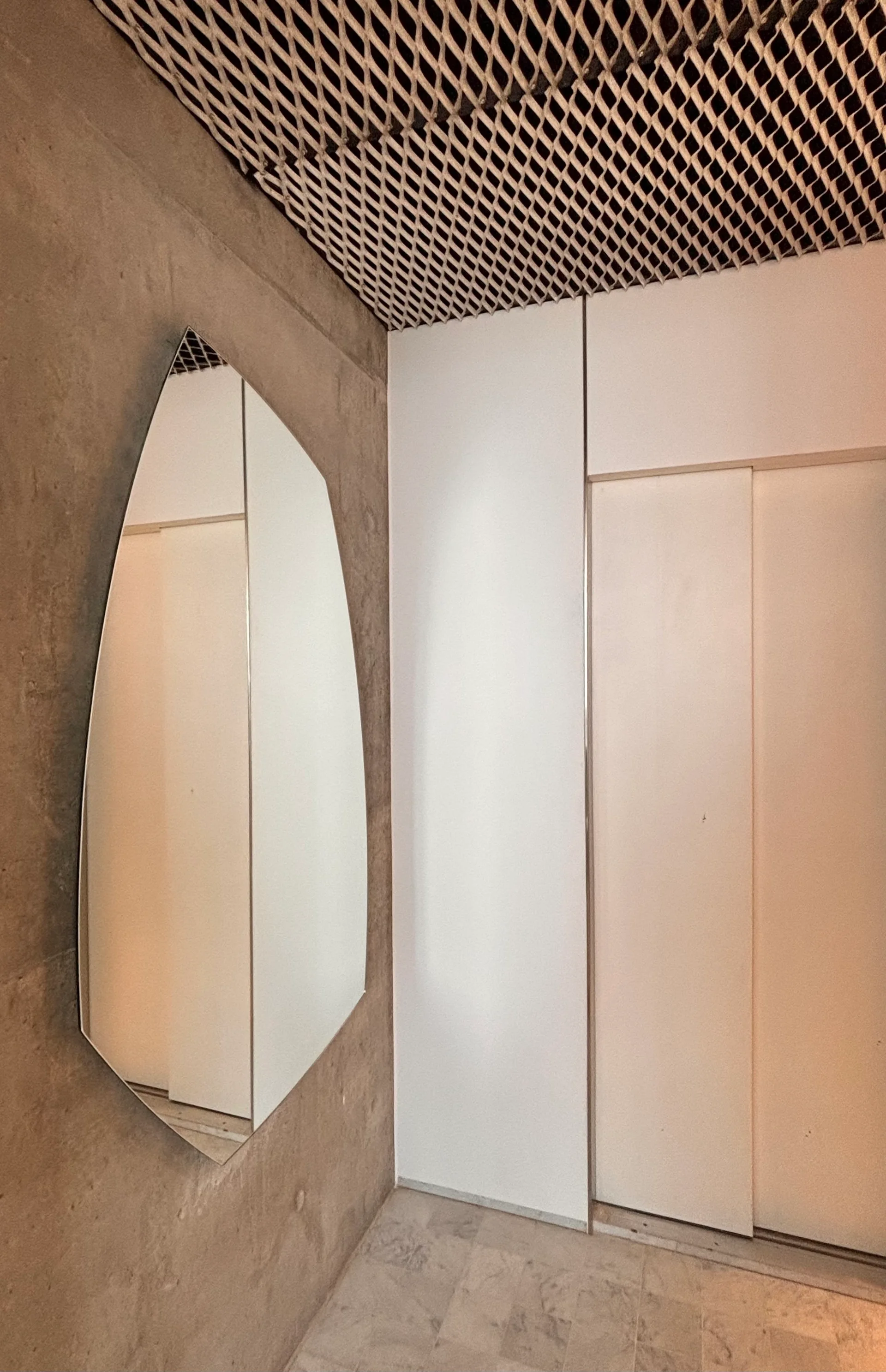 An asymmetrical geometric mirror hangs on a textured concrete wall next to flush white closet doors under a metal mesh ceiling.
