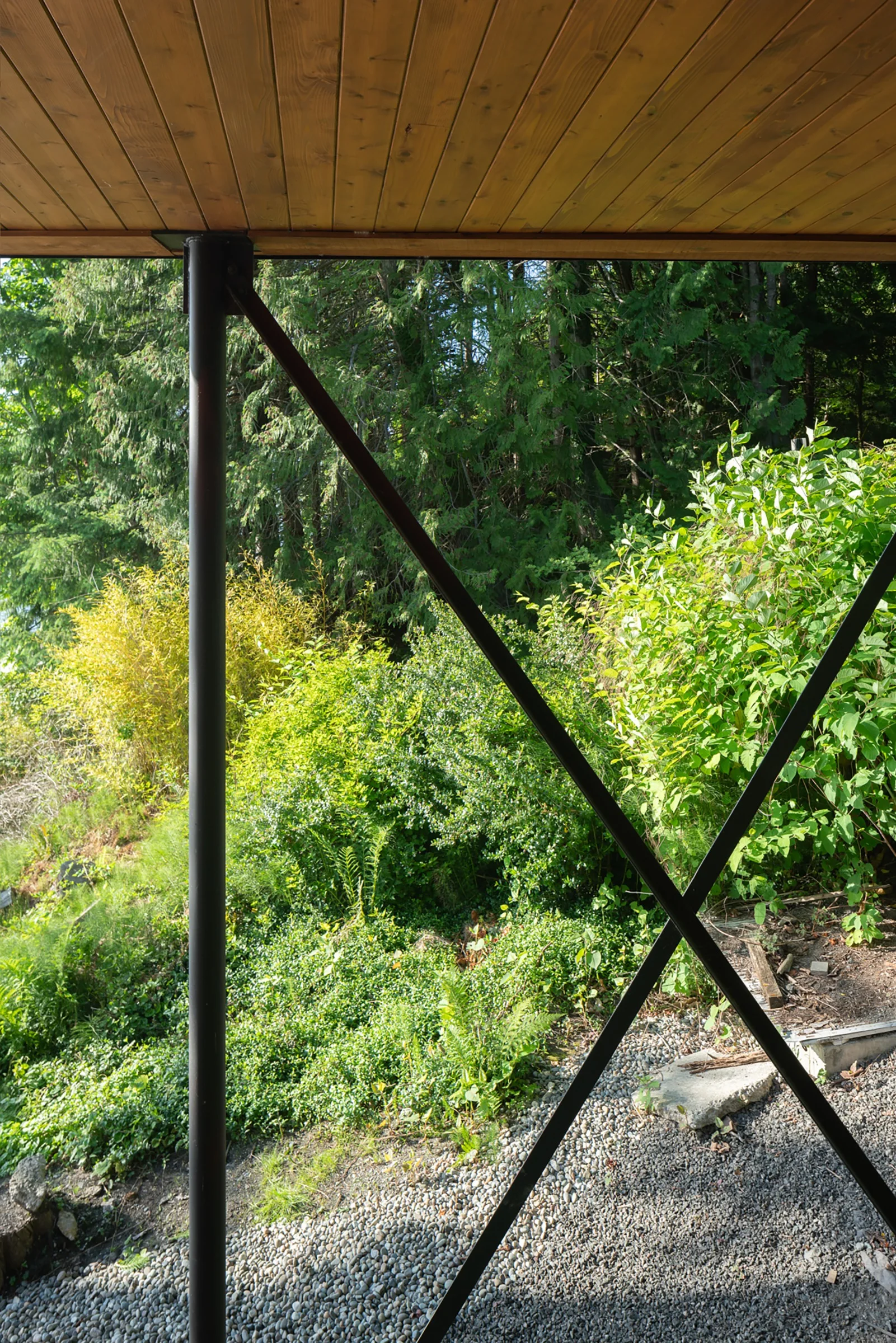 A covered porch features a polished wood plank ceiling, black metal support posts with cross-bracing, and a view of a lush green forest.