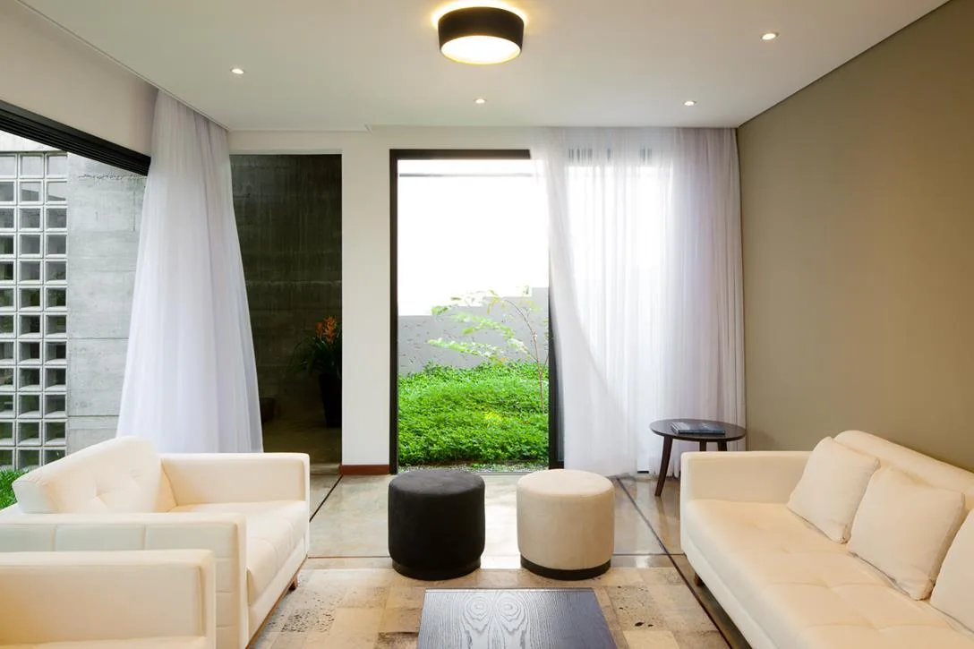 White sofas and round ottomans face each other in a bright living room next to large glass doors with sheer white curtains opening to a garden.