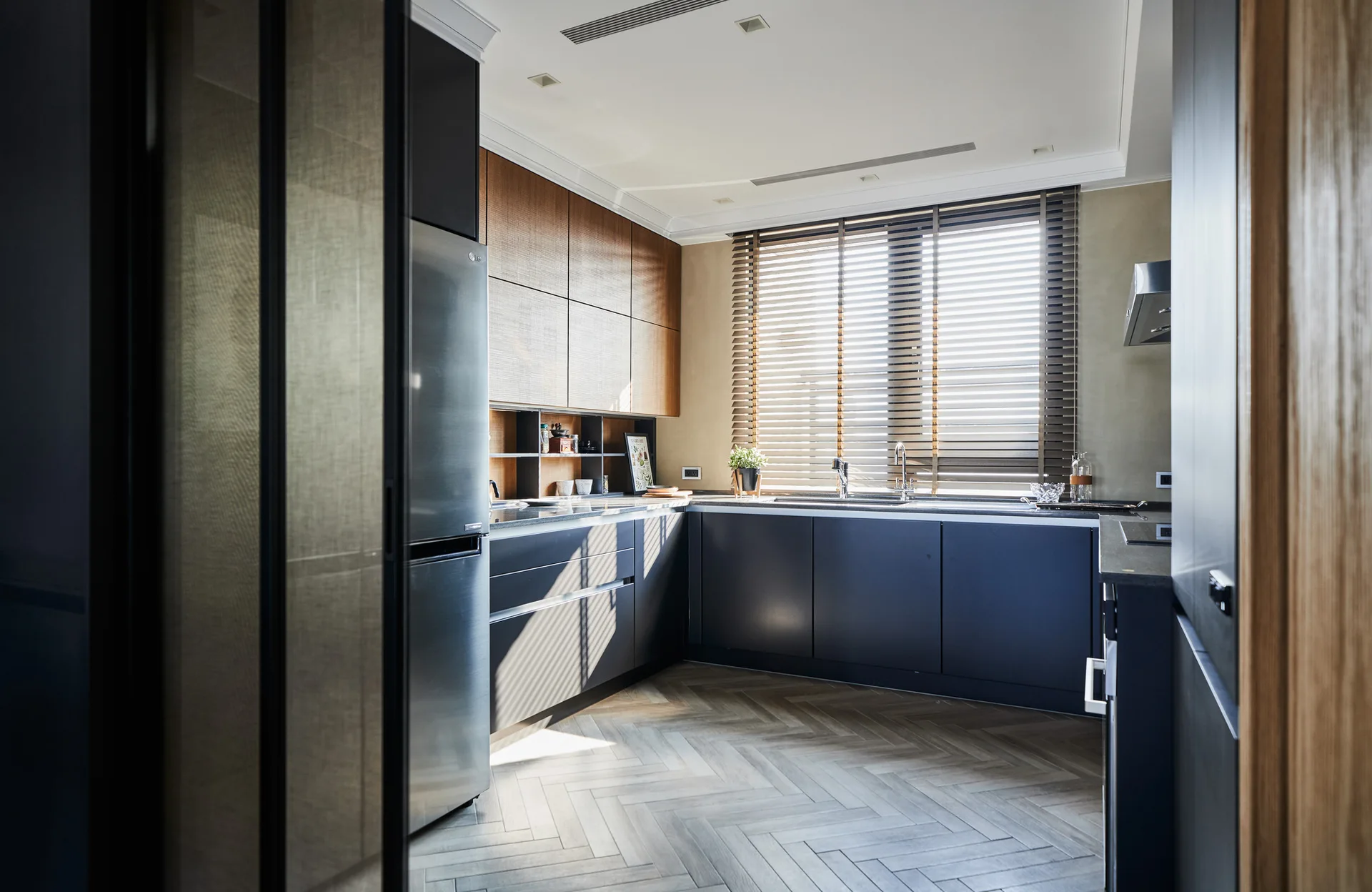 A modern kitchen featuring two-tone charcoal and wood cabinets, a stainless steel refrigerator, herringbone flooring, and horizontal window blinds.