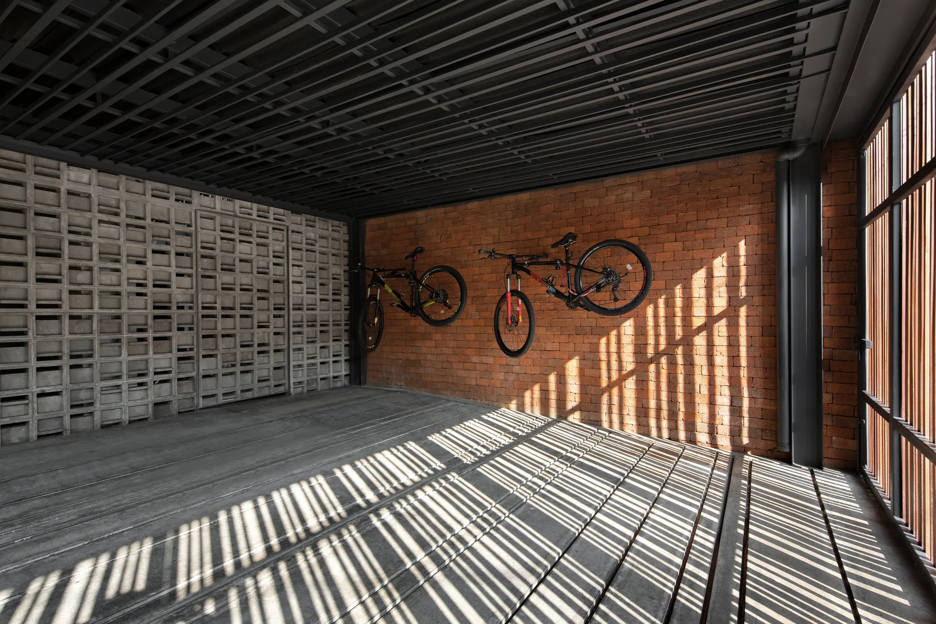 Two mountain bikes hang on an exposed red brick wall inside a modern garage featuring a concrete breezeblock partition and slatted shadows.