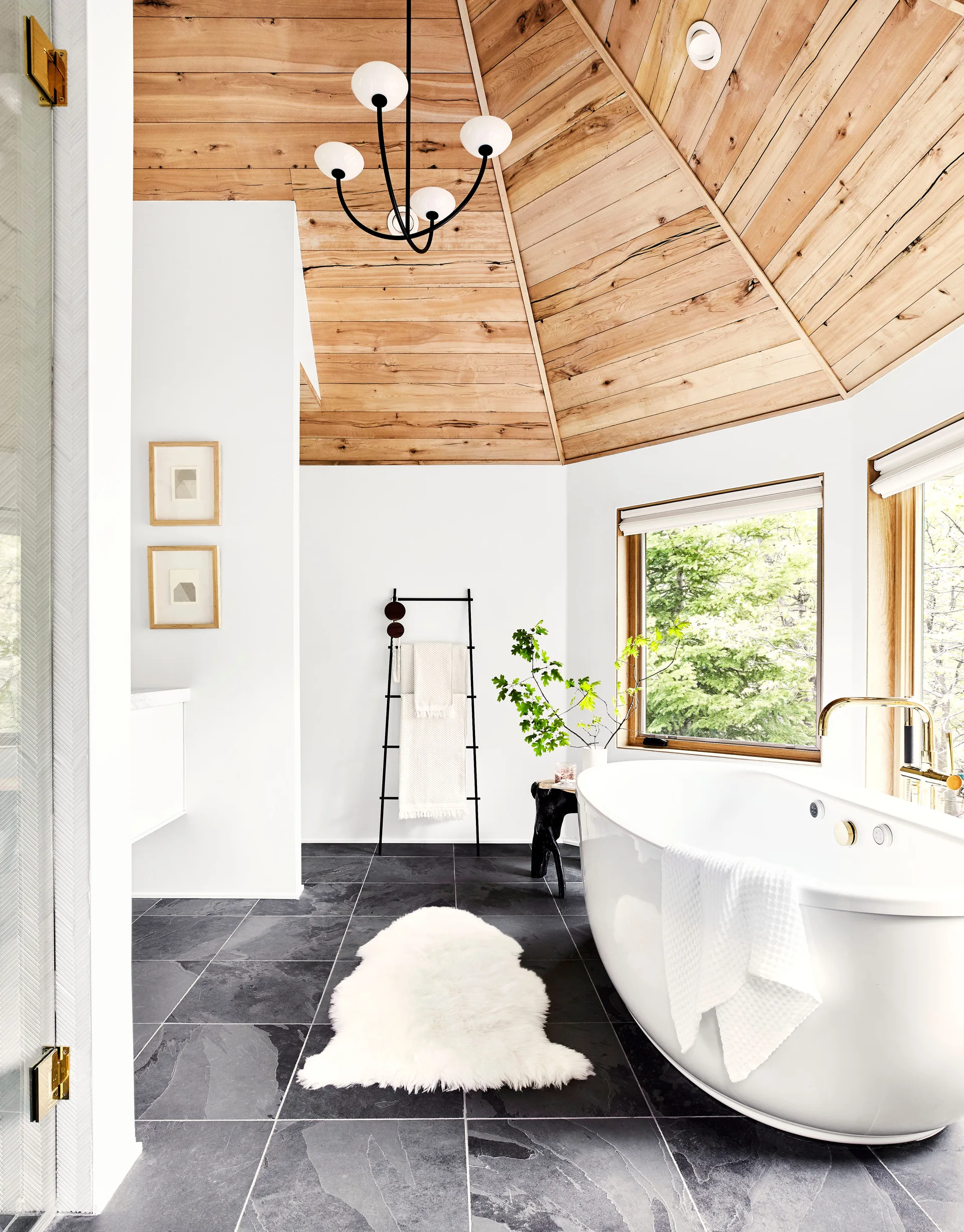A modern bathroom features a white freestanding bathtub on a dark slate tile floor beneath a vaulted wood-plank ceiling.