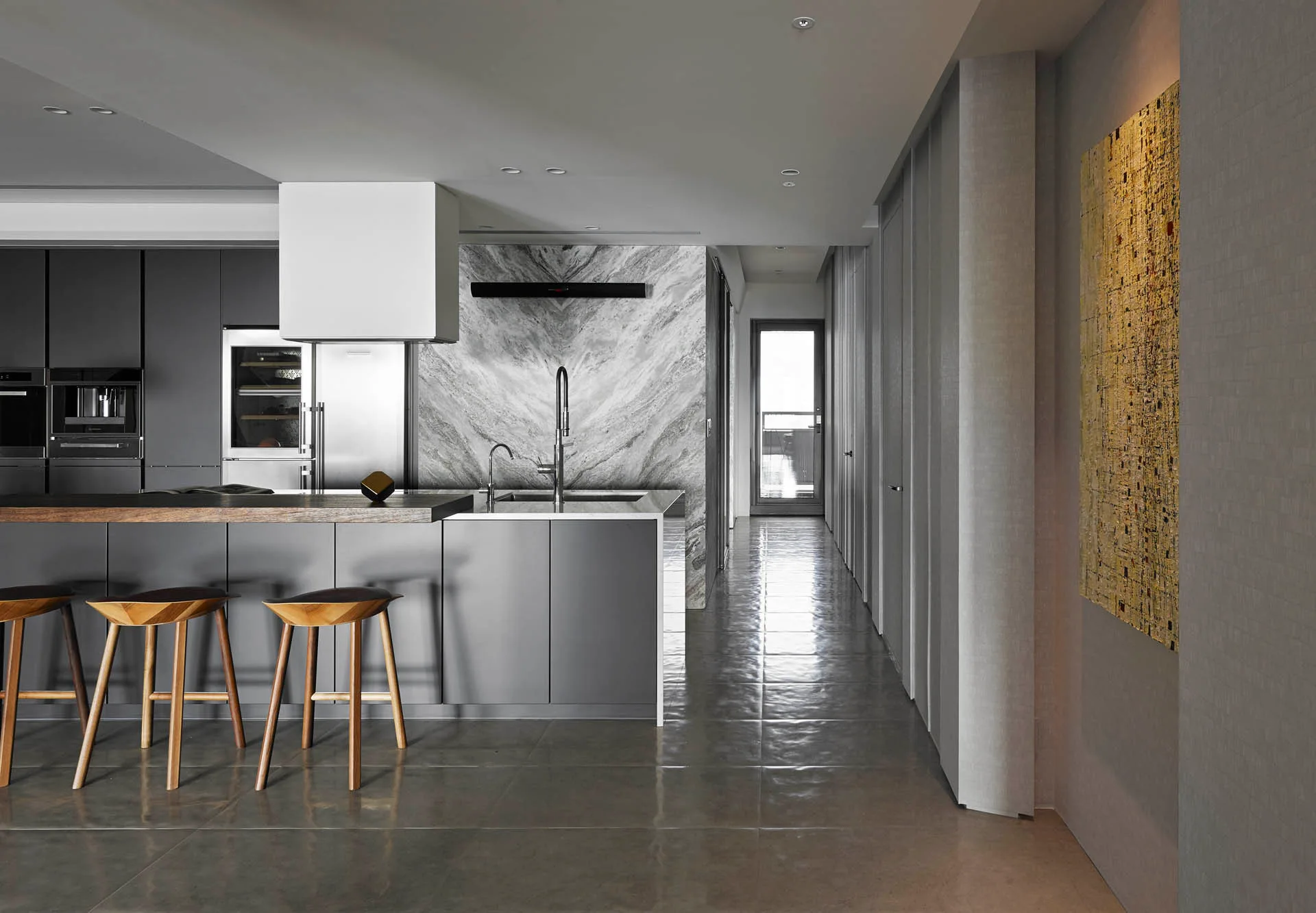 A minimalist modern kitchen with a charcoal gray island, three wooden bar stools, and a highly veined marble backsplash.