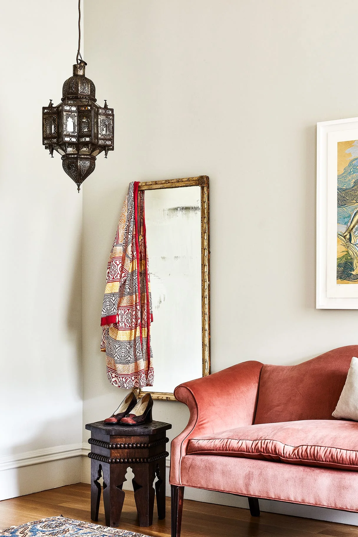 A living room corner features a pink velvet sofa beside a gilded floor mirror, a carved wooden side table, and an intricate metal pendant lantern.