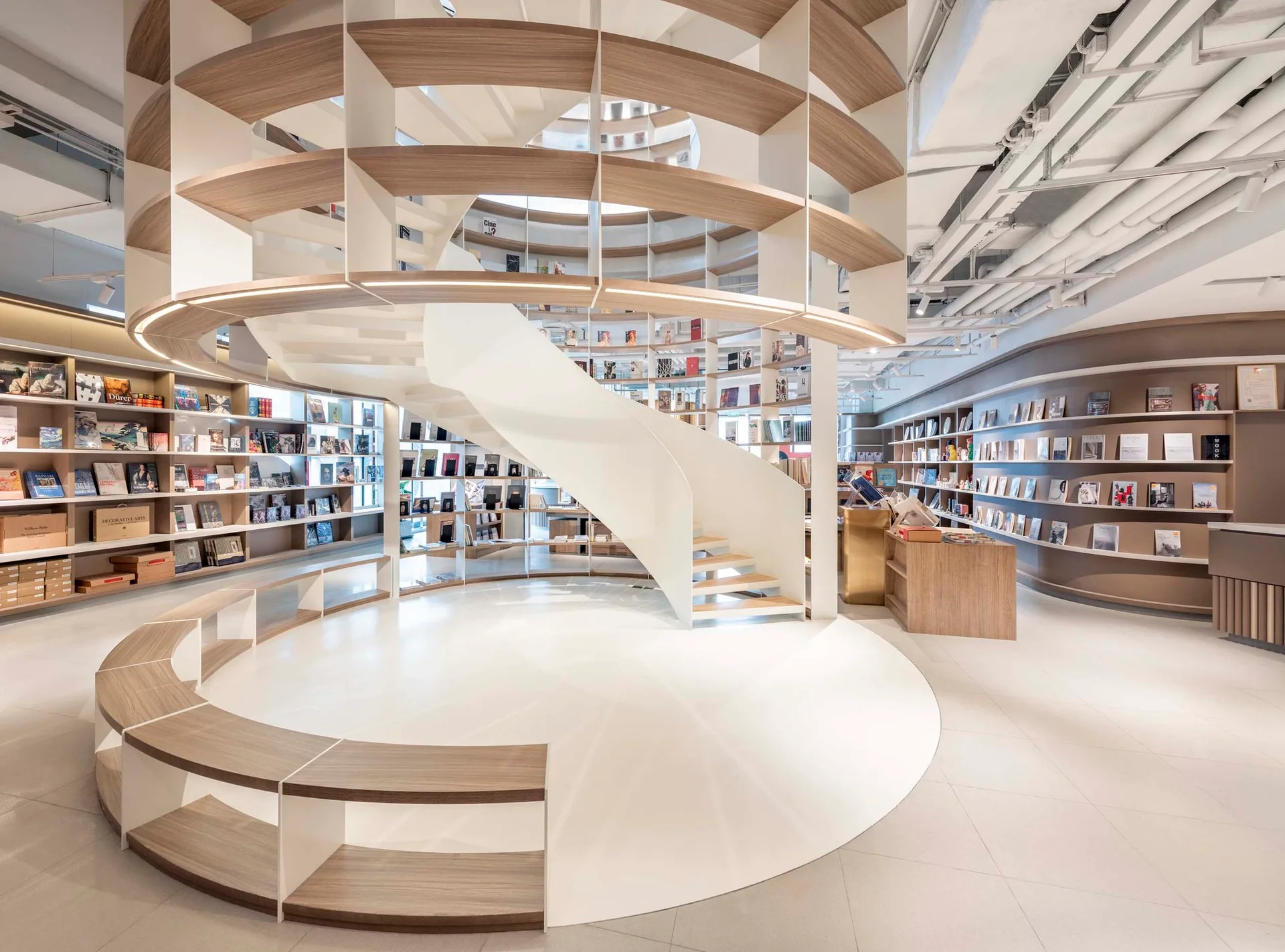 A modern bookstore interior featuring a central white spiral staircase surrounded by curved wooden shelving units and matching circular bench seating.