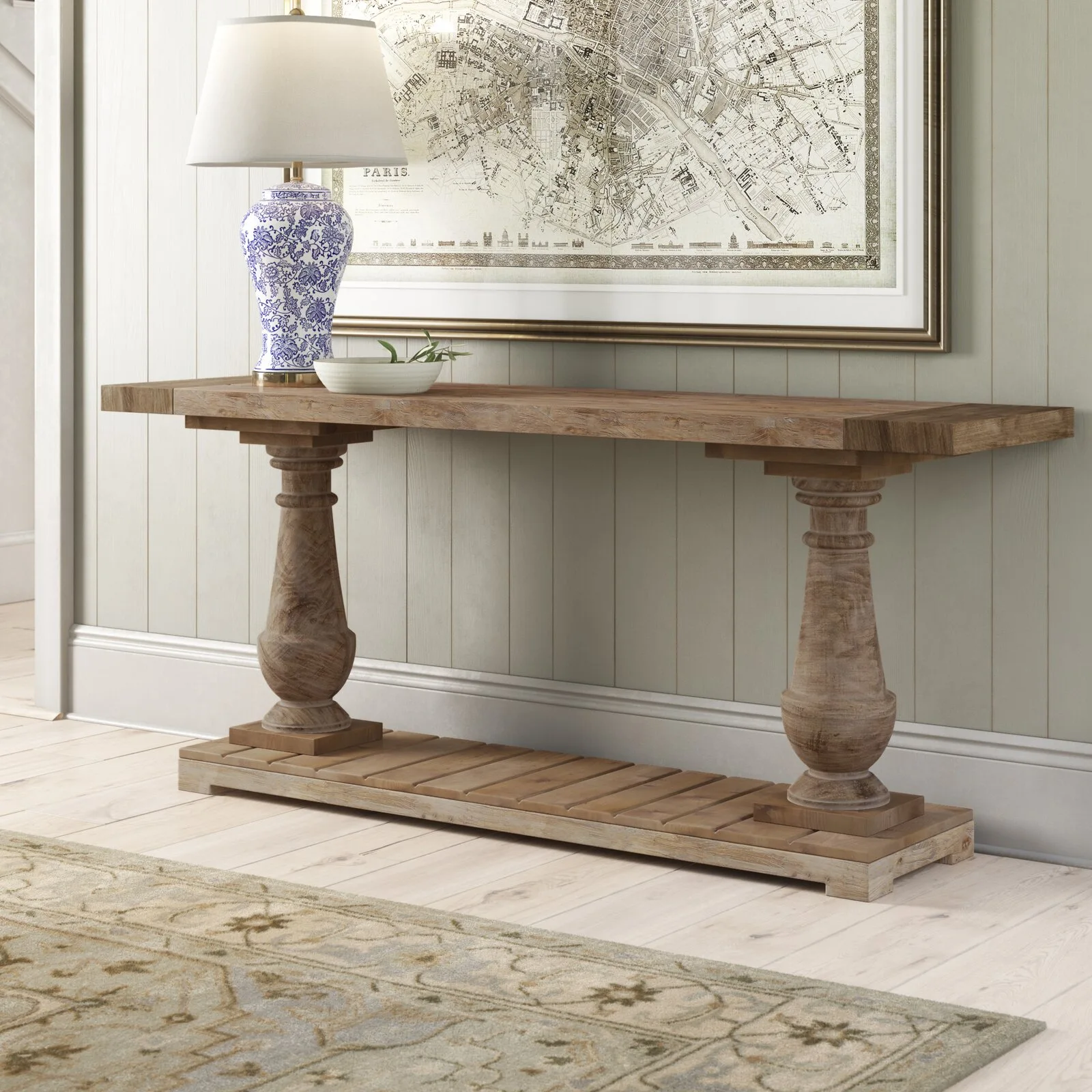 A rustic wooden console table with turned legs is set against a vertical paneled wall, decorated with a blue and white patterned lamp and a framed map.