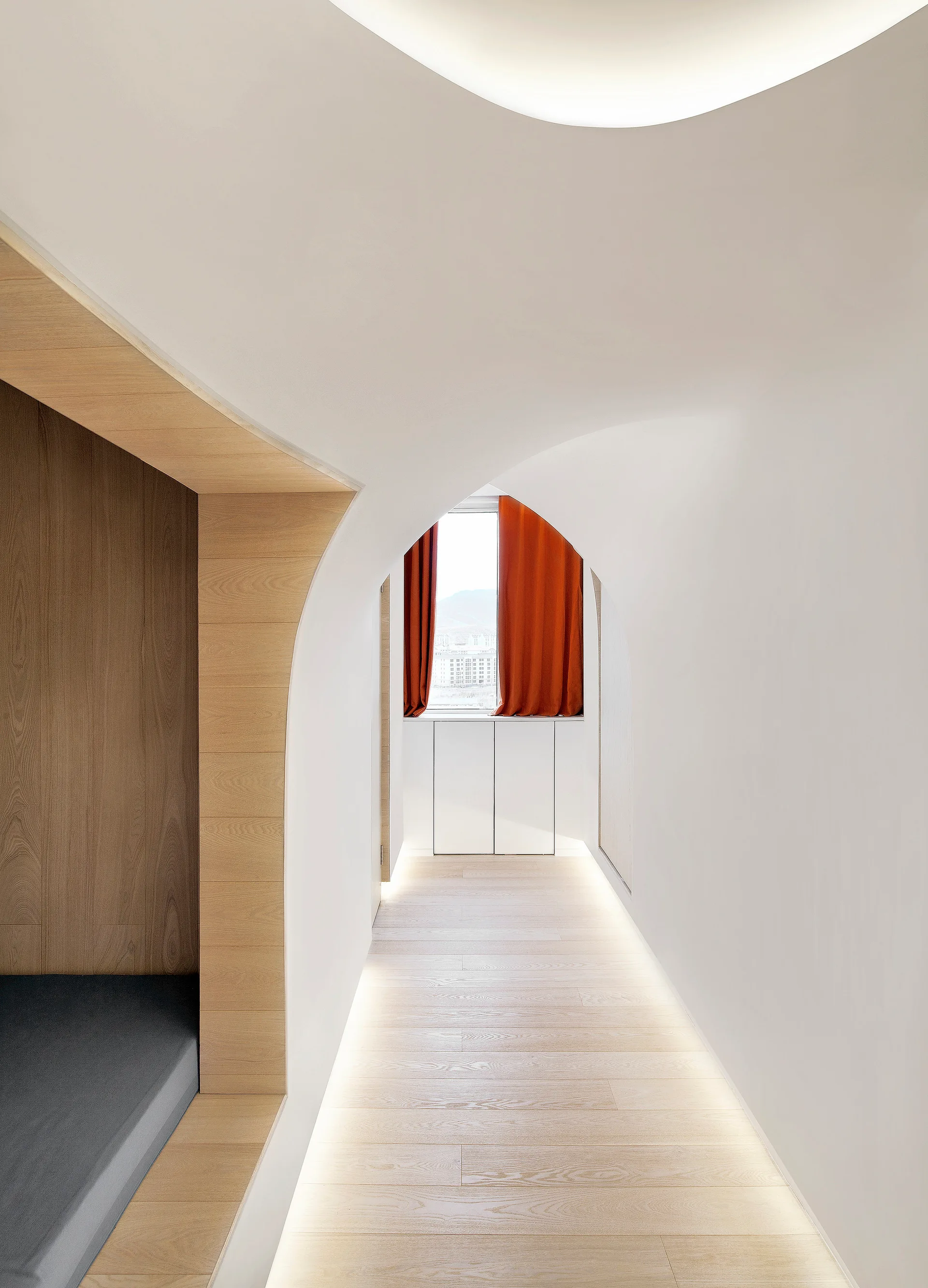 A minimalist white hallway features an arched passage, light wood flooring with recessed baseboard lighting, and a window with orange curtains.