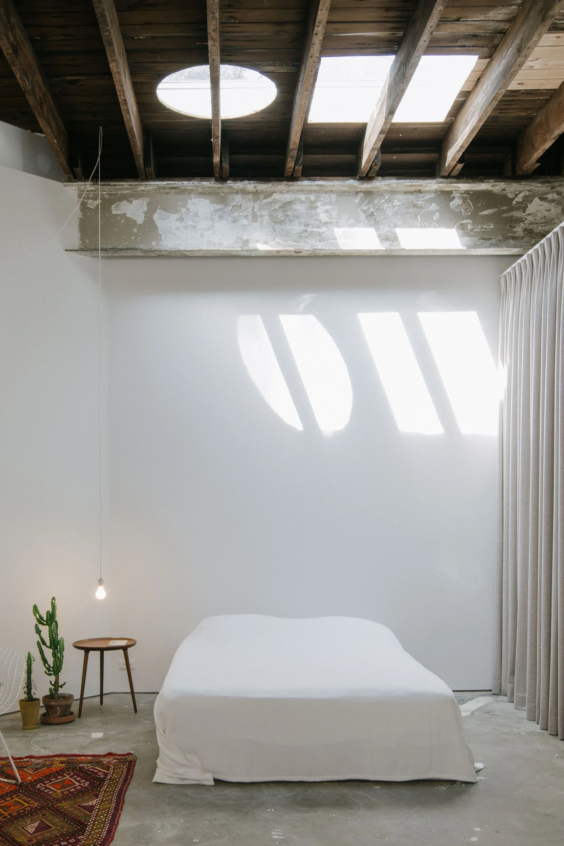 A minimalist loft bedroom features a white bed on a concrete floor beneath an exposed wooden rafter ceiling with dual skylights.