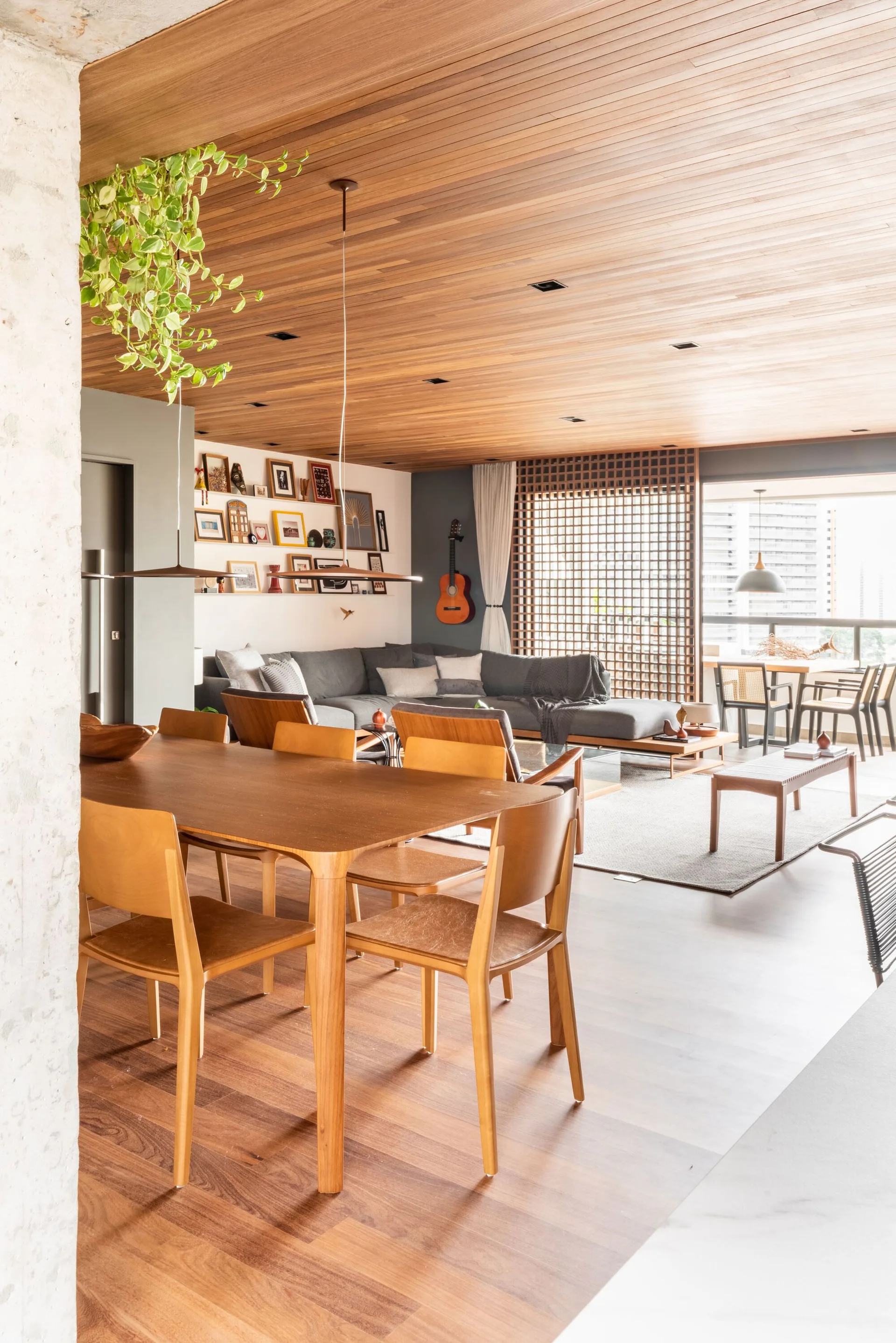 An open-concept living space features a wooden slatted ceiling, a mid-century dining set, a grey sectional sofa, and a geometric wood lattice window screen.