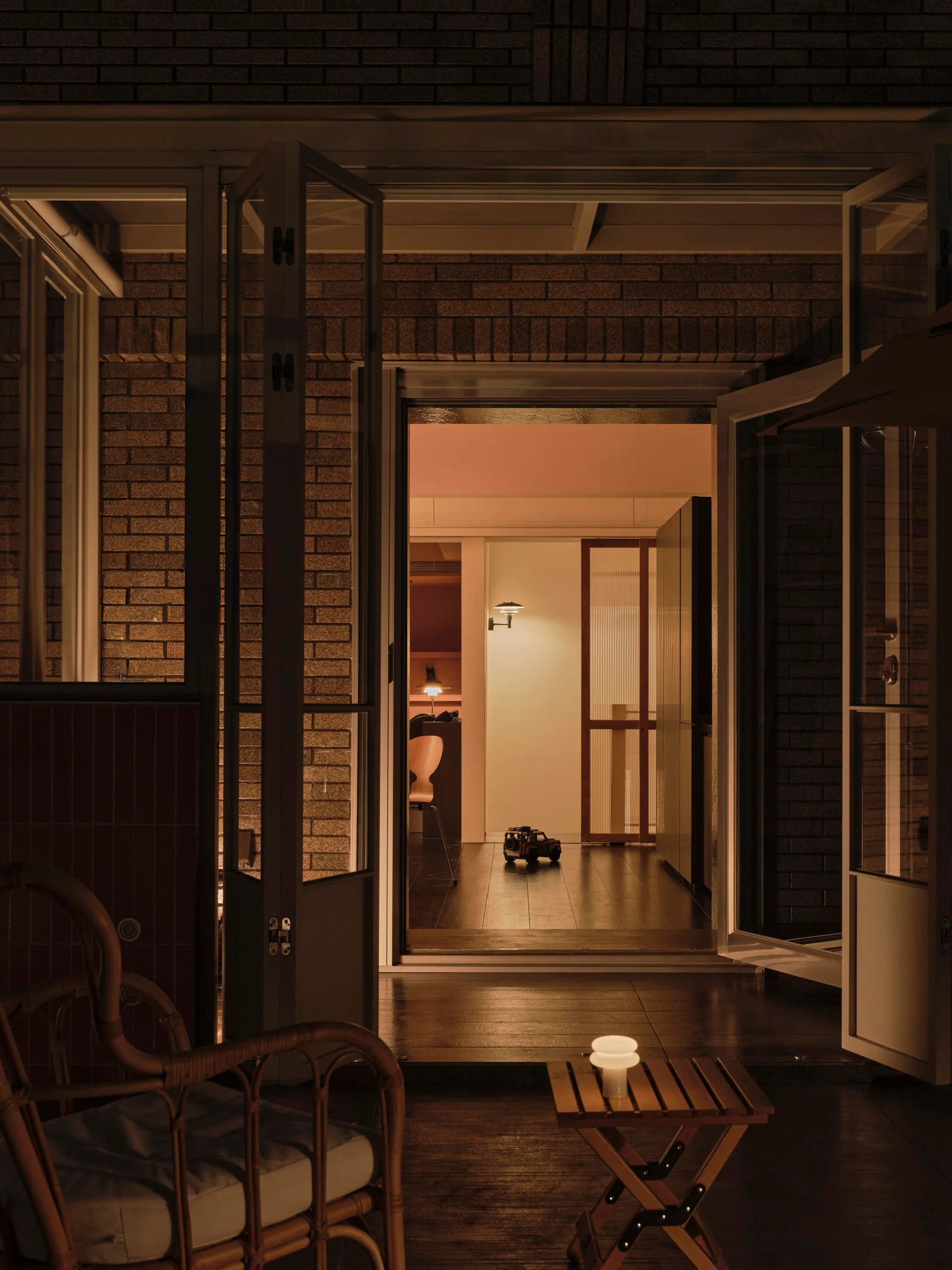 An open set of glass doors leads from a brick-walled terrace to a warmly lit interior room featuring a fluted glass screen.