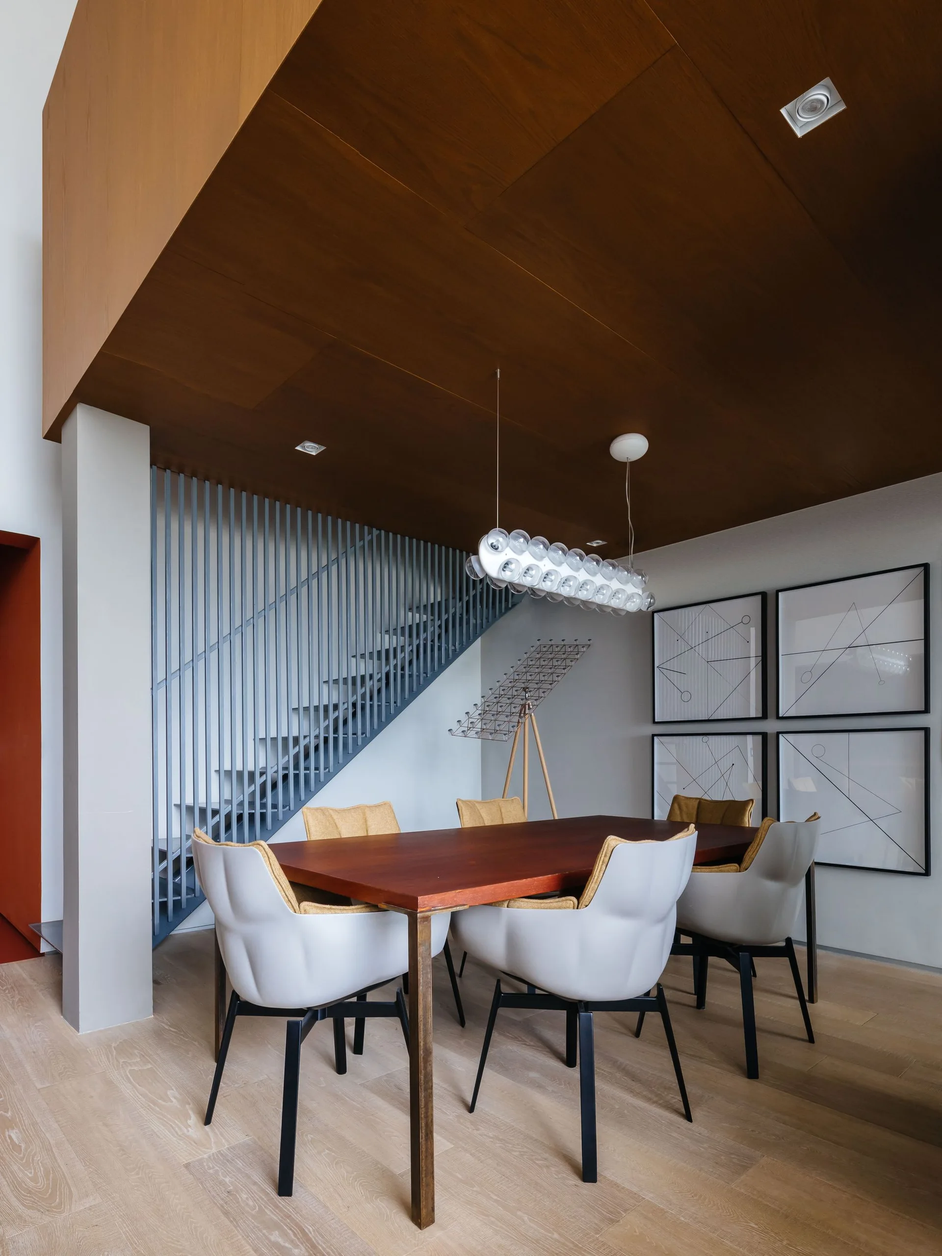 A modern dining room features a dark wood table with white chairs beneath a wood-paneled ceiling canopy next to an open staircase.